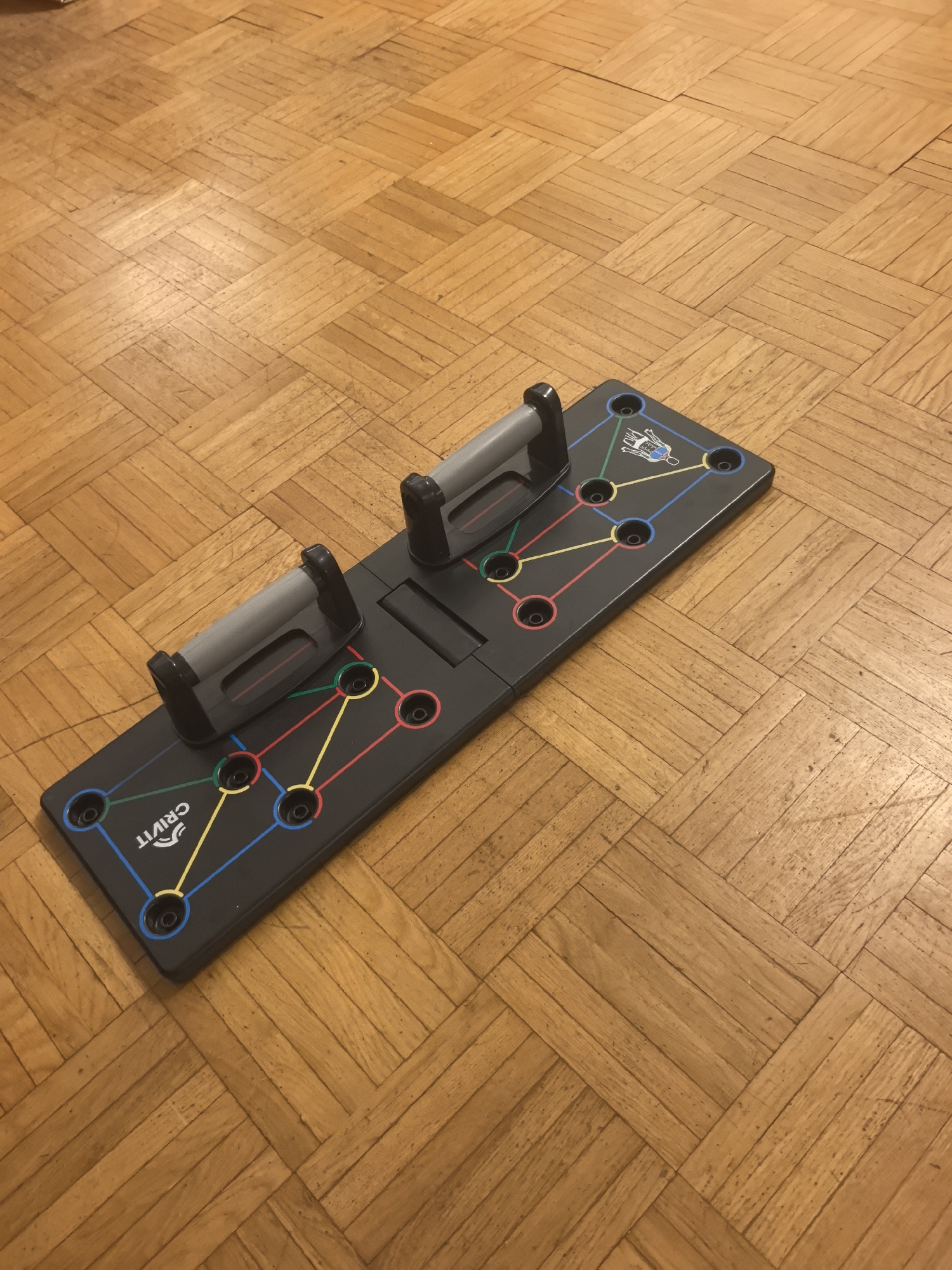 The image features a black, foldable push-up board with two detachable grey handles, placed on a light-colored wooden parquet floor. The floor has a distinct herringbone pattern and shows some minor marks or dust from use.
The push-up board is designed with multiple circular holes, each outlined with a specific color (red, yellow, green, or blue). These holes are connected by corresponding colored lines, indicating different positions for the handles to target various muscle groups. A small white diagram of a human figure is printed on the board, illustrating the muscle areas associated with the handle placements. The handles themselves are grey with black bases and are currently inserted into two of the designated holes. The brand name "CRIVIT" is visible in white text on the left side of the board.
The scene is indoors, likely a room within a residence in Hattersheim am Main, Germany. The lighting appears to be ambient, typical of an interior setting. No activity is depicted; the equipment is static, set up and ready for use.