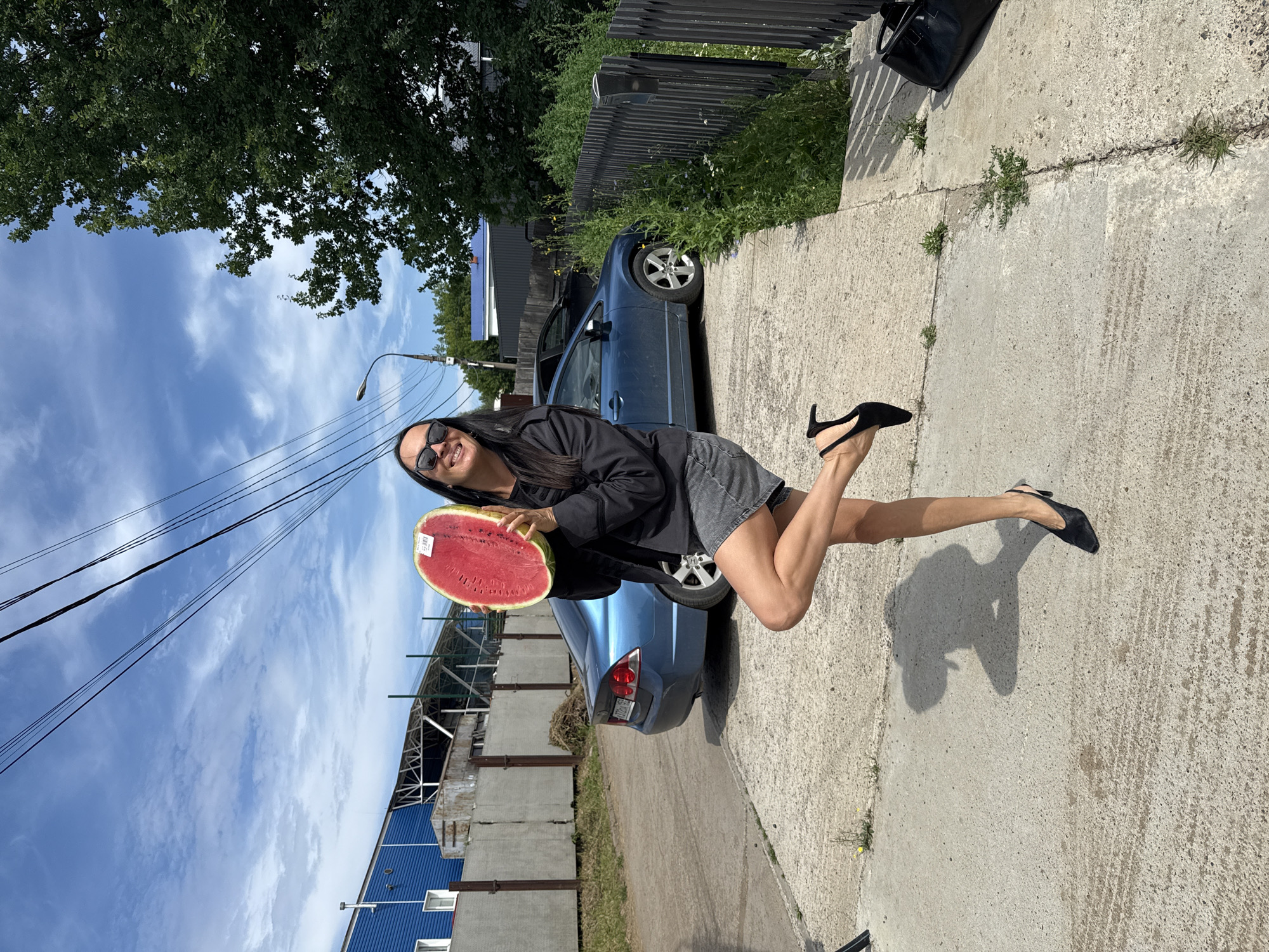 In an outdoor, urban environment consistent with Chaikovsky, Russia, a woman stands on a concrete path, holding a large slice of watermelon above her head. She is positioned to the left of a blue sedan, which is parked on a dirt and gravel area behind her. The woman is wearing a black zip-up hoodie, a denim skirt, and black high-heeled shoes. She is also wearing sunglasses and has long, dark hair. Her left leg is bent at the knee, with her foot raised off the ground. Behind the car, a black wooden fence runs along the right side of the frame, bordering an area with tall green grass and weeds. In the background, a blue industrial building with a metal roof and a white fence is visible, along with power lines stretched across the sky. The sky is blue with scattered white clouds. A black bag is placed on the ground near the fence on the right side of the image.