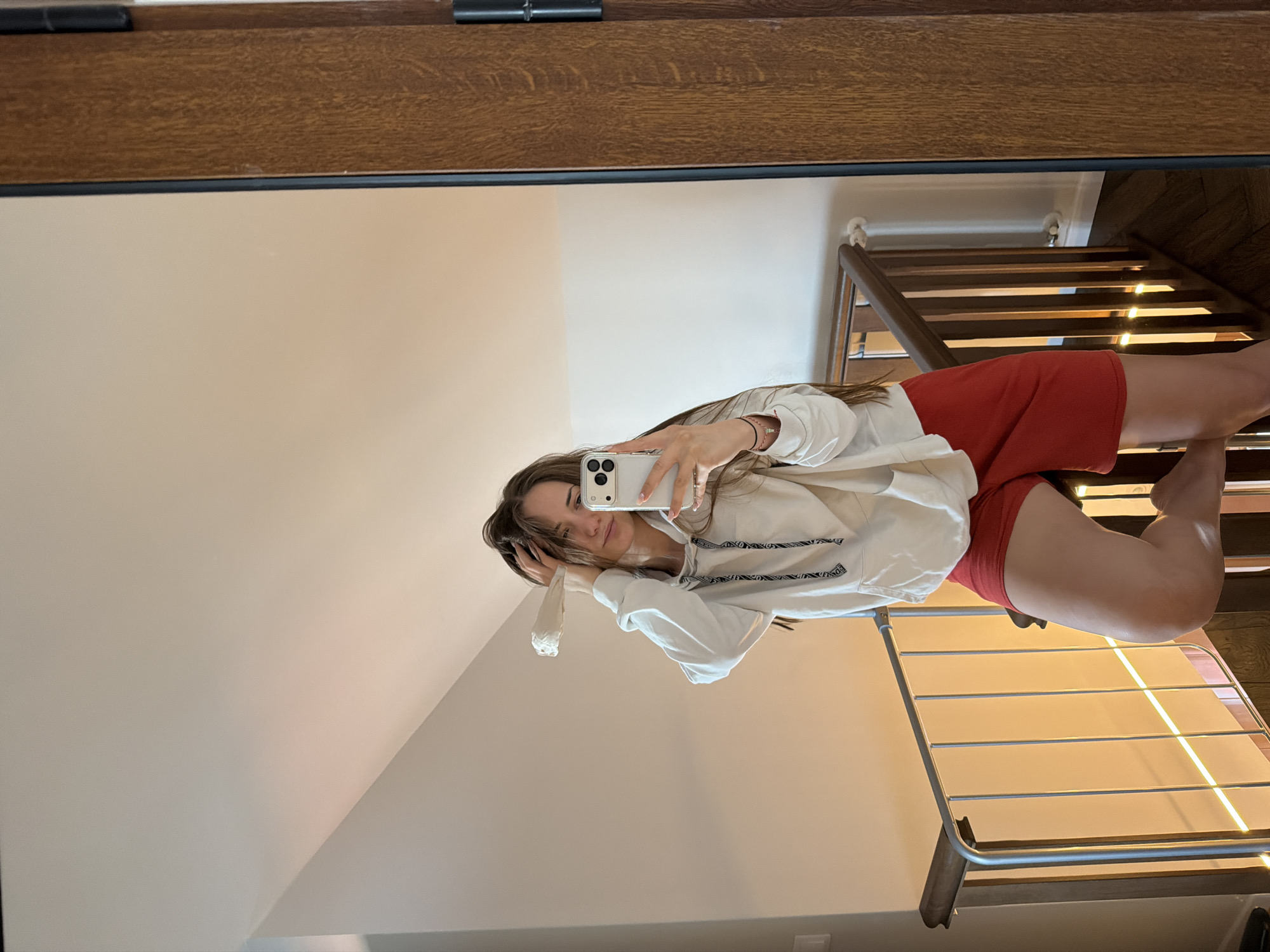 A young woman is reflected in a mirror. She holds a white smartphone in her right hand, presumably taking a selfie. She wears a light-colored hooded sweatshirt and red shorts. A staircase is visible behind her in the reflection. The environment appears to be indoors, likely a home. There is no indication of location in the city of Buhovo, Bulgaria from the image.