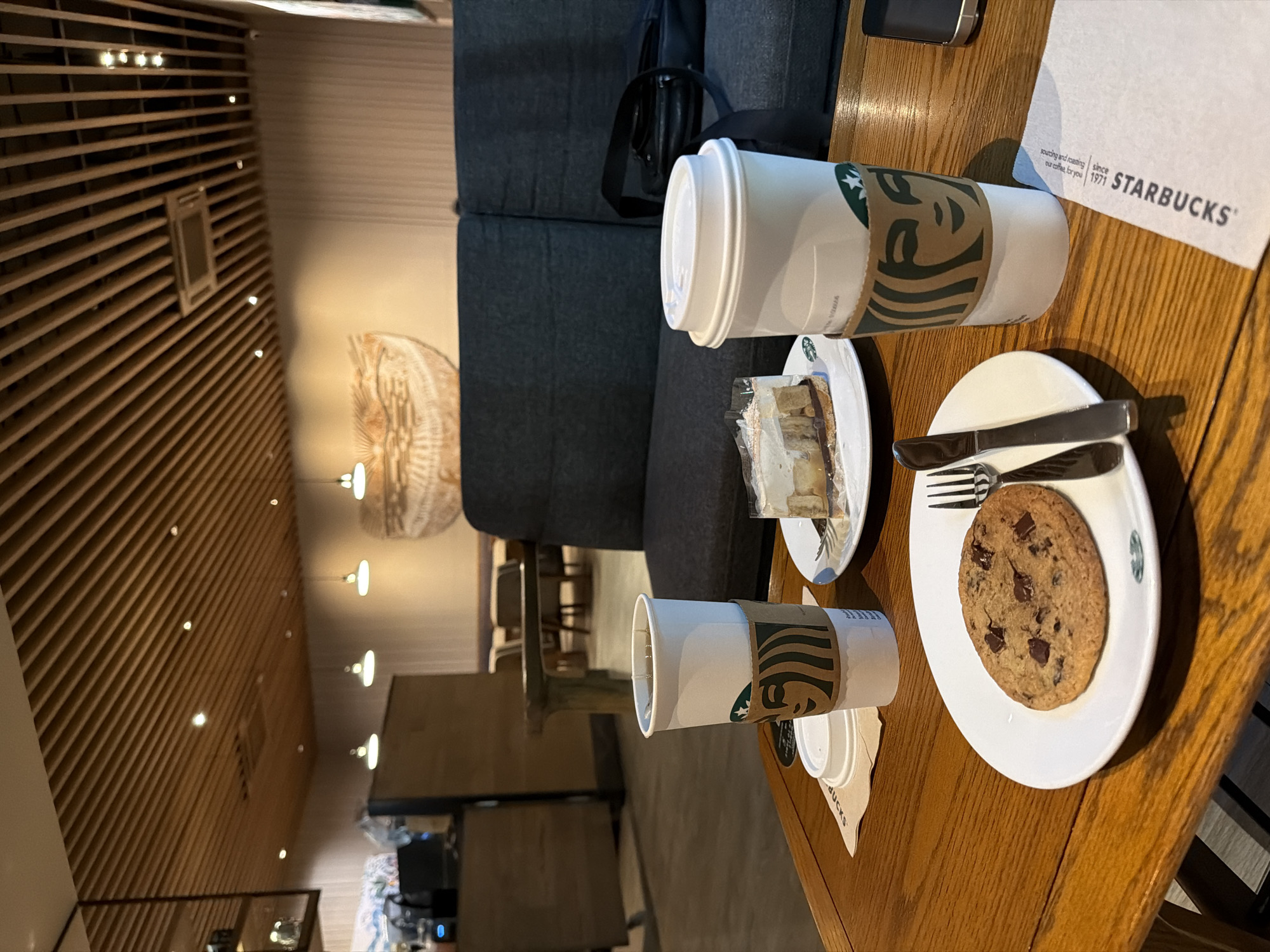 The image shows a table at a Starbucks in Pattaya City, Thailand. On the table are a cookie, a piece of cake, two Starbucks drinks in paper cups, and cutlery on a white plate. The environment includes a sofa, a wooden table, and interior lighting with decorative wooden beams on the ceiling. There are no people visible in the frame.