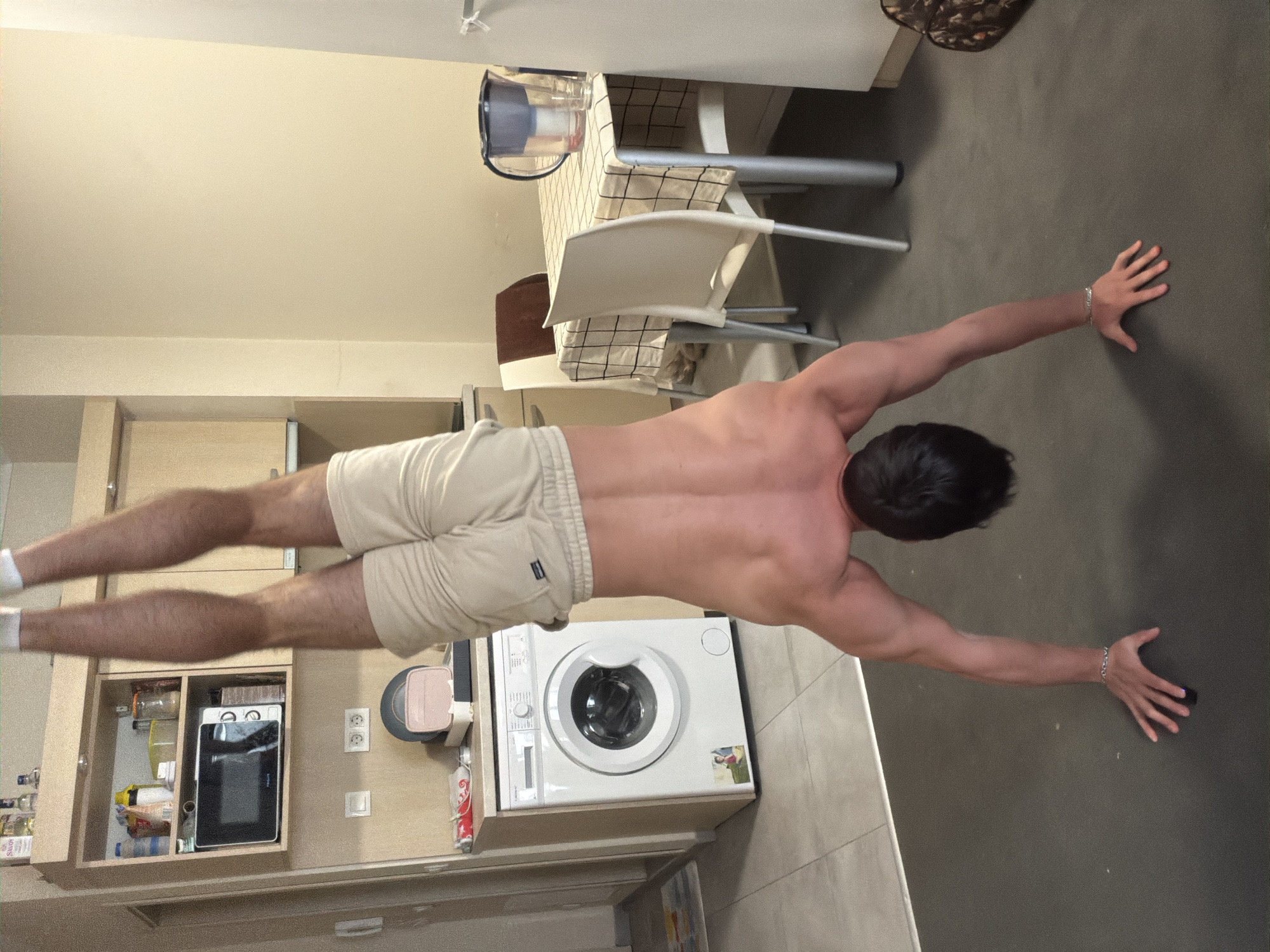 A shirtless man with a muscular physique, including a well-defined back and arms, performs a horizontal, plank-like exercise. He is positioned with his body parallel to the floor, stretching his arms forward and legs backward, supported by his hands flat on the dark grey floor. He wears light beige athletic shorts and white socks, with a silver bracelet on his left wrist.
The scene is an indoor domestic setting, likely an apartment in Sofia, Bulgaria, combining a kitchen area with a living or dining space. To the man's left, there is a light wood-toned kitchen counter featuring open shelving containing a microwave oven and various food items and bottles. Electrical outlets are visible on the wall above the counter. Directly beneath the man's torso and legs is a white front-loading washing machine.
To the man's right, a dining table with a white and black checkered tablecloth is set with a clear glass pitcher. Two modern white chairs with silver frames are positioned around the table. The walls are a light beige, and the ceiling is white. The overall lighting is even, consistent with an indoor daytime setting. No significant text is clearly legible on any visible objects. The man's pose indicates concentration and physical exertion.
