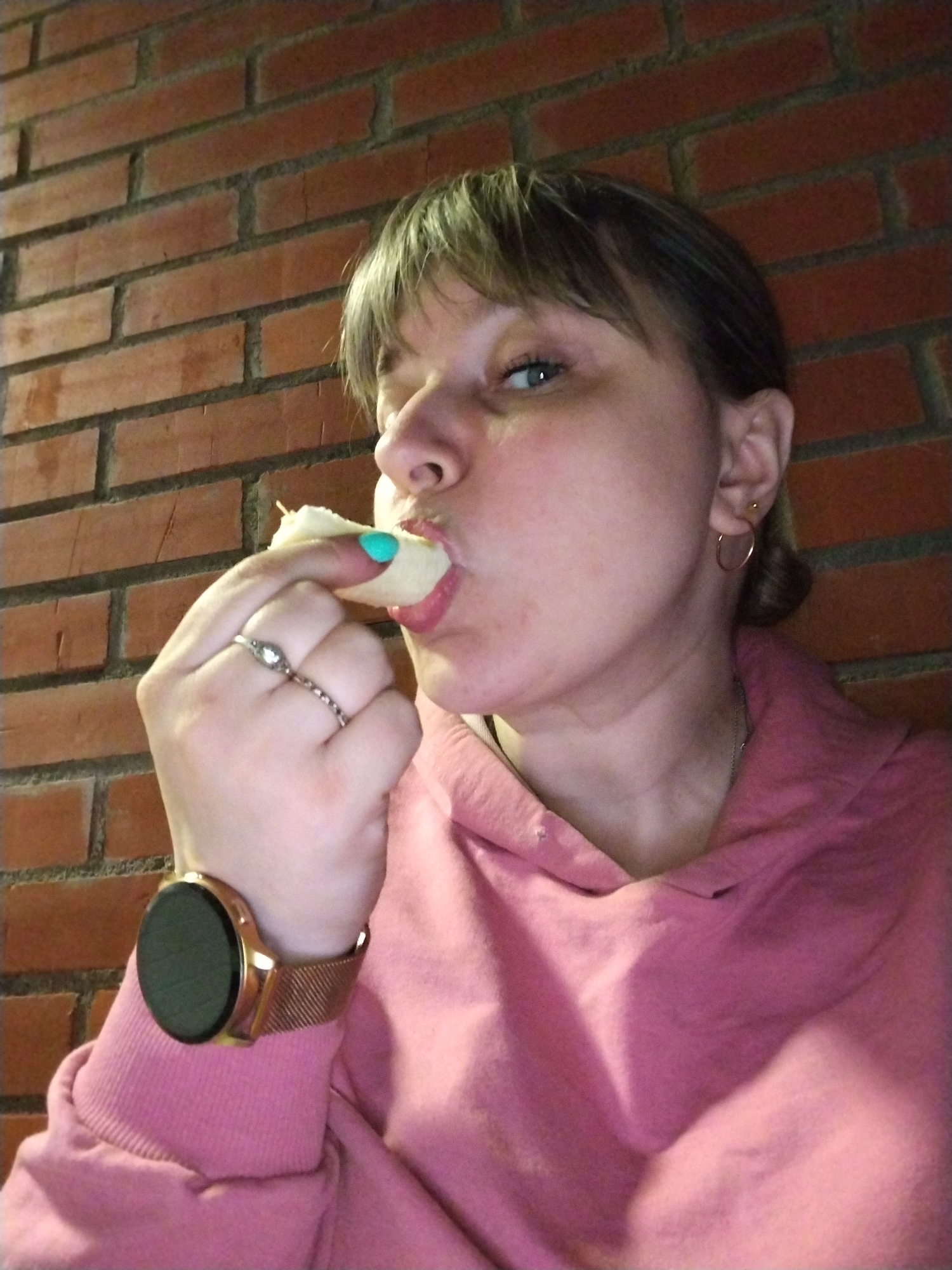A woman is captured in a close-up, profile shot, eating a light-colored food item. She is positioned against a brick wall, suggesting an indoor setting. Her mouth is open, biting into the food, which she holds with her right hand.
The woman has light brown hair with bangs. She wears a pink hoodie or sweatshirt. Notable accessories include a ring with a clear stone on her middle finger and a rose gold-toned smartwatch with a mesh strap on her left wrist. She also has small, gold hoop earrings. Her right thumb, holding the food, features bright turquoise or teal nail polish. Her eyes are looking slightly upward and to the right, with a focused expression as she eats.
The background is a reddish-brown brick wall, providing a textured backdrop. No text is visible in the image. The lighting appears to be artificial, casting some shadows but illuminating the subject well.