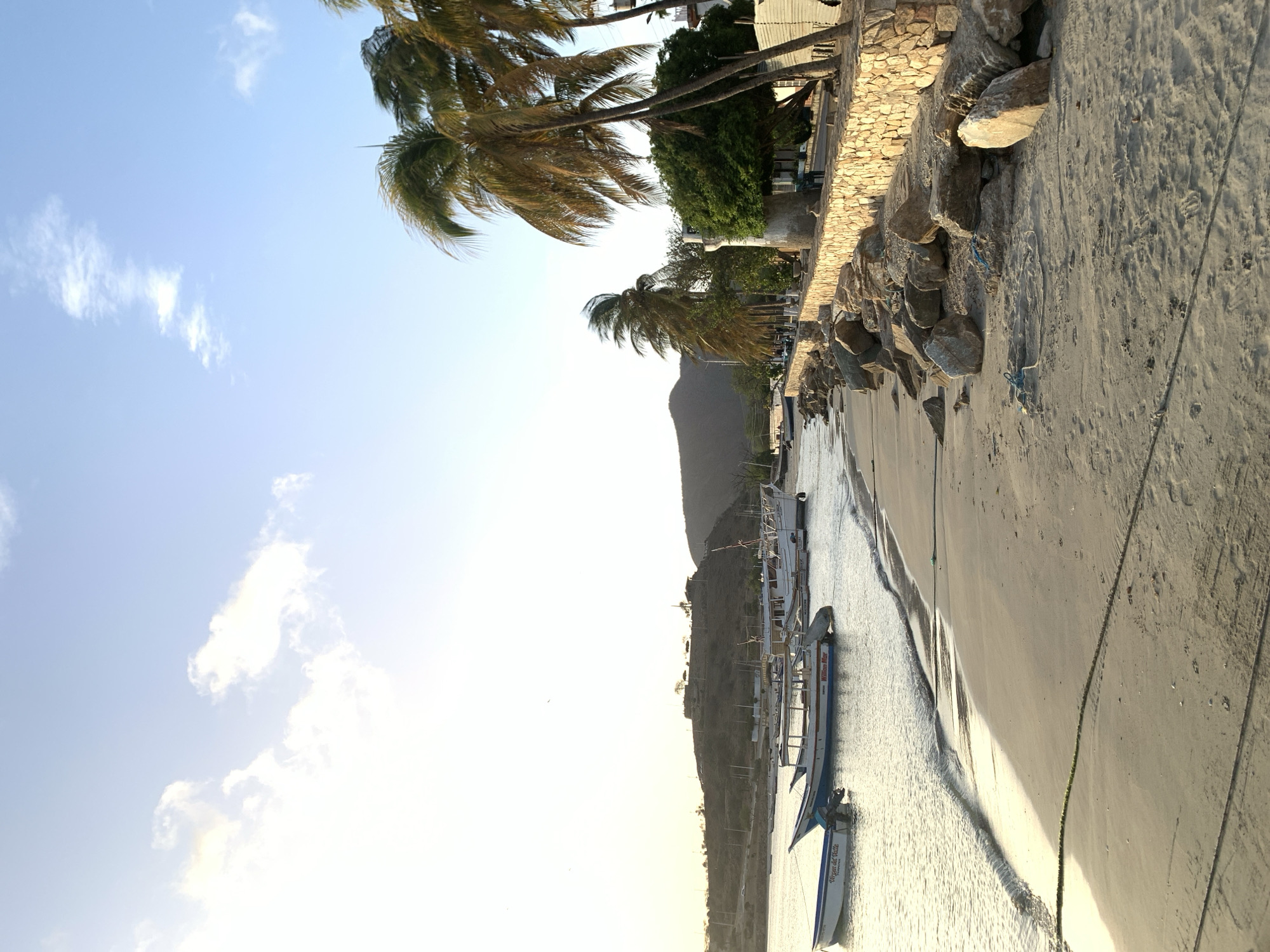 The image depicts a scenic view of the waterfront in Juan Griego, Venezuela, at what appears to be either sunrise or sunset. The foreground is dominated by a sandy beach with a few scattered rocks and some mooring ropes. Gentle waves lap at the shore.
In the water, several boats are anchored. The closest boat on the left has "Virgen del Valle" written on its side. Further out, a larger white boat with red trim is visible, along with other smaller vessels.
A stone wall lines the promenade behind the beach, and several tall palm trees sway in the breeze. Beyond the promenade, there are buildings and a lush green tree. In the background, a hilly landscape rises, with some structures visible on its crest. The sky is a clear blue with some wispy clouds, illuminated by the soft light of the sun. No people are clearly visible in the image.