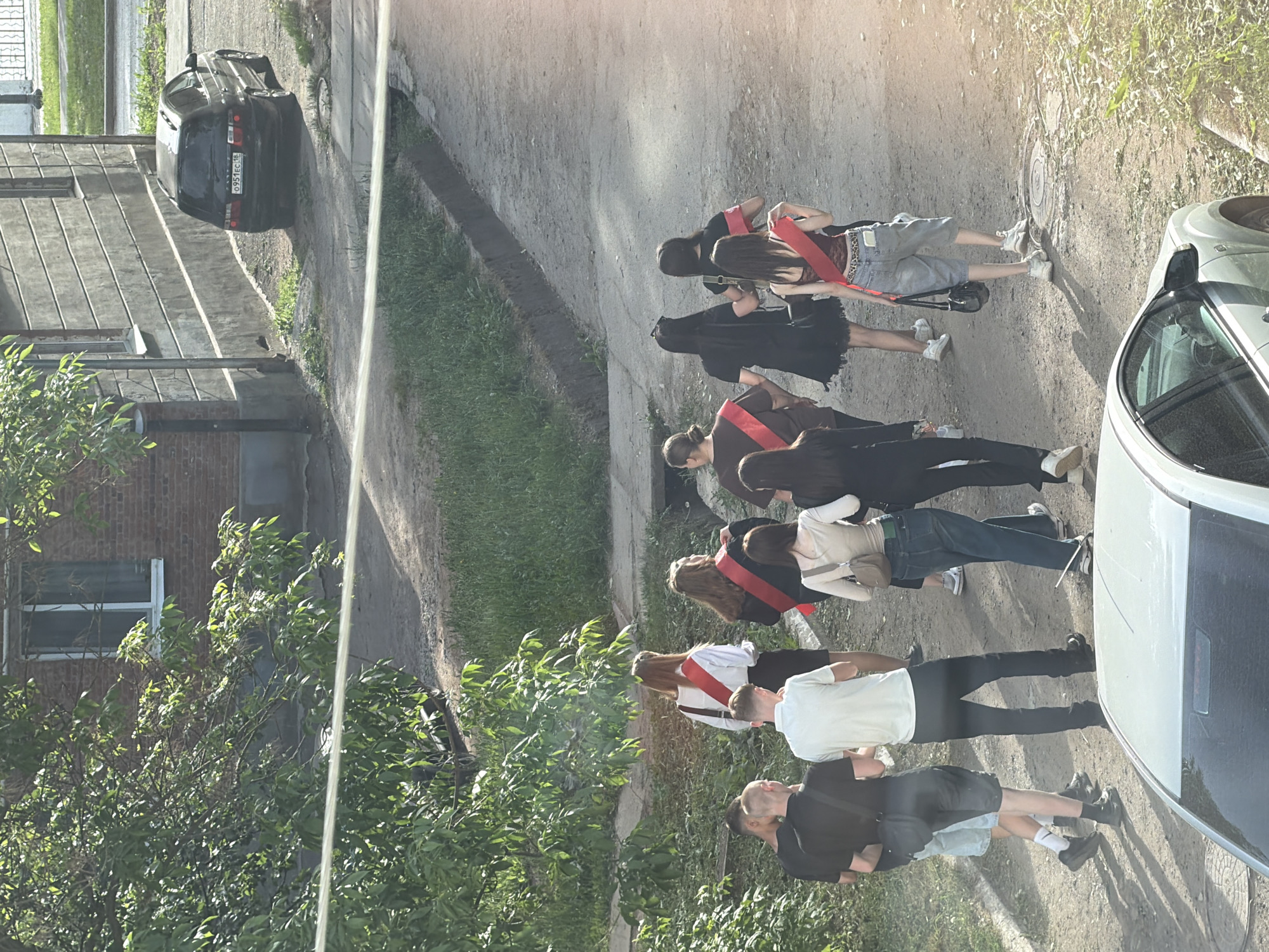 A medium-shot photograph captures a group of people, appearing to be graduates, walking in a line along a street in Votkinsk, Russia. The graduates are walking on a partially paved road beside a grassy area and a sidewalk. They are dressed in a mix of formal and casual clothing, with some wearing red sashes indicating graduation.
Several individuals are visible: The front two people wear black shirts with gray shorts. Other people wear black or white shirts with jeans. Some are wearing dresses. All the people are wearing shoes and carrying bags or purses.
A white car is parked on the right, partially obscuring the view. On the left, there is a brick building, a metal fence, and another car parked. The license plate is partly visible. The setting appears to be a residential area, possibly near an educational institution.