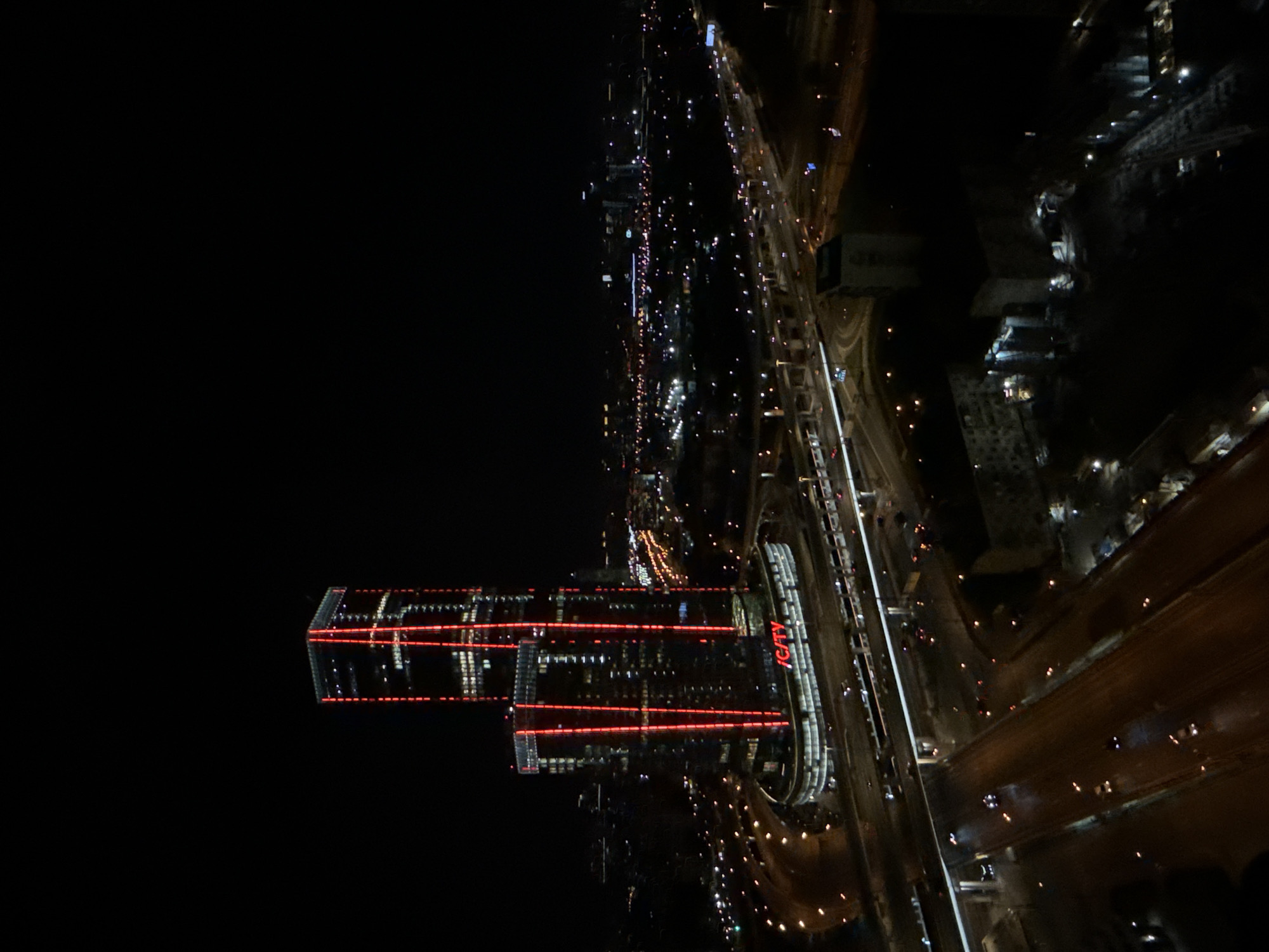 The image displays a nighttime aerial or elevated view of an urban environment, likely in Khimki, Russia. The original image is rotated 90 degrees clockwise; when viewed upright, it presents a dynamic cityscape illuminated by artificial lights against a dark sky.
In the left portion of the upright frame, a prominent cluster of modern, high-rise buildings stands tall. These architectural structures feature extensive red illumination, with distinct horizontal and vertical red light strips tracing their glass facades. One of the most noticeable buildings clearly displays the word "iCITY" in vertical red lettering along its side, serving as a key identifying landmark within the scene.
To the right and extending across the rest of the upright image, a vast and intricate network of highways and roads stretches into the distance. These thoroughfares are bustling with vehicle activity, evidenced by countless streaks and dots of white headlights and red taillights, indicating significant traffic flow. Elevated sections and multiple lanes of the highway system are visible, weaving through the dark urban landscape.
The time of day is unequivocally night, with no natural light present. The sky appears clear, suggesting good visibility and allowing for a sharp view of the brightly lit cityscape. There are no visible indications of adverse weather conditions. The scene captures the grandeur and constant motion of a vibrant, modern metropolitan area, active even under the cover of darkness, with artificial lighting effectively highlighting its architectural and infrastructural elements.