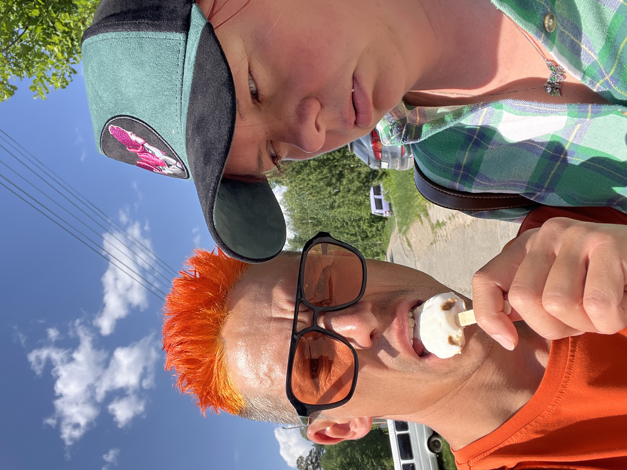 Two people pose for a photo outdoors on a sunny day with blue skies and white clouds. One person, wearing a green plaid shirt and a butterfly necklace, is positioned above and slightly behind the other. The second person has bright orange hair and is wearing black-framed sunglasses and an orange shirt. This person is holding and eating a white ice cream bar. The location appears to be in Chaikovsky, Russia, as inferred from previous context. Visible in the background is a road, green trees, a white van, and a light purple vehicle.