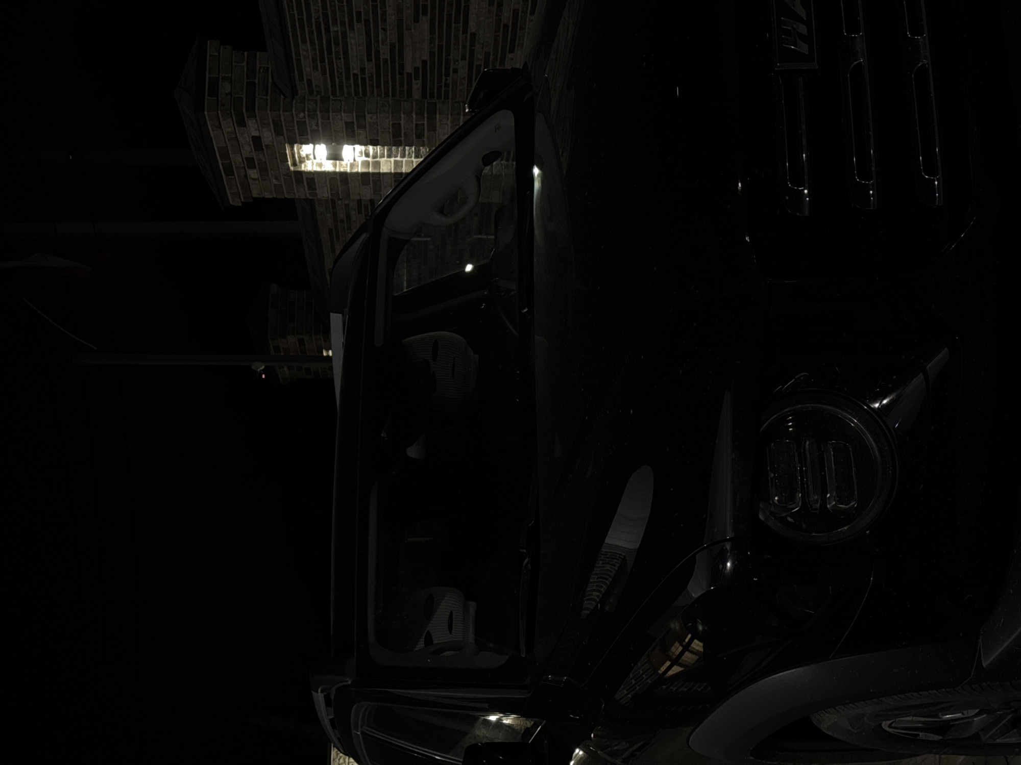 A low-angle, partially-lit photograph shows the front passenger side of a black Hummer vehicle, parked inside a garage or covered area. The vehicle’s passenger door is open, and the interior of the vehicle is dimly visible. The Hummer's front grille is visible on the right. The upper part of the scene contains a brick-lined ceiling, which is partially illuminated by a light fixture. The photograph's environment is dark, and the majority of the image is obscured by shadows.