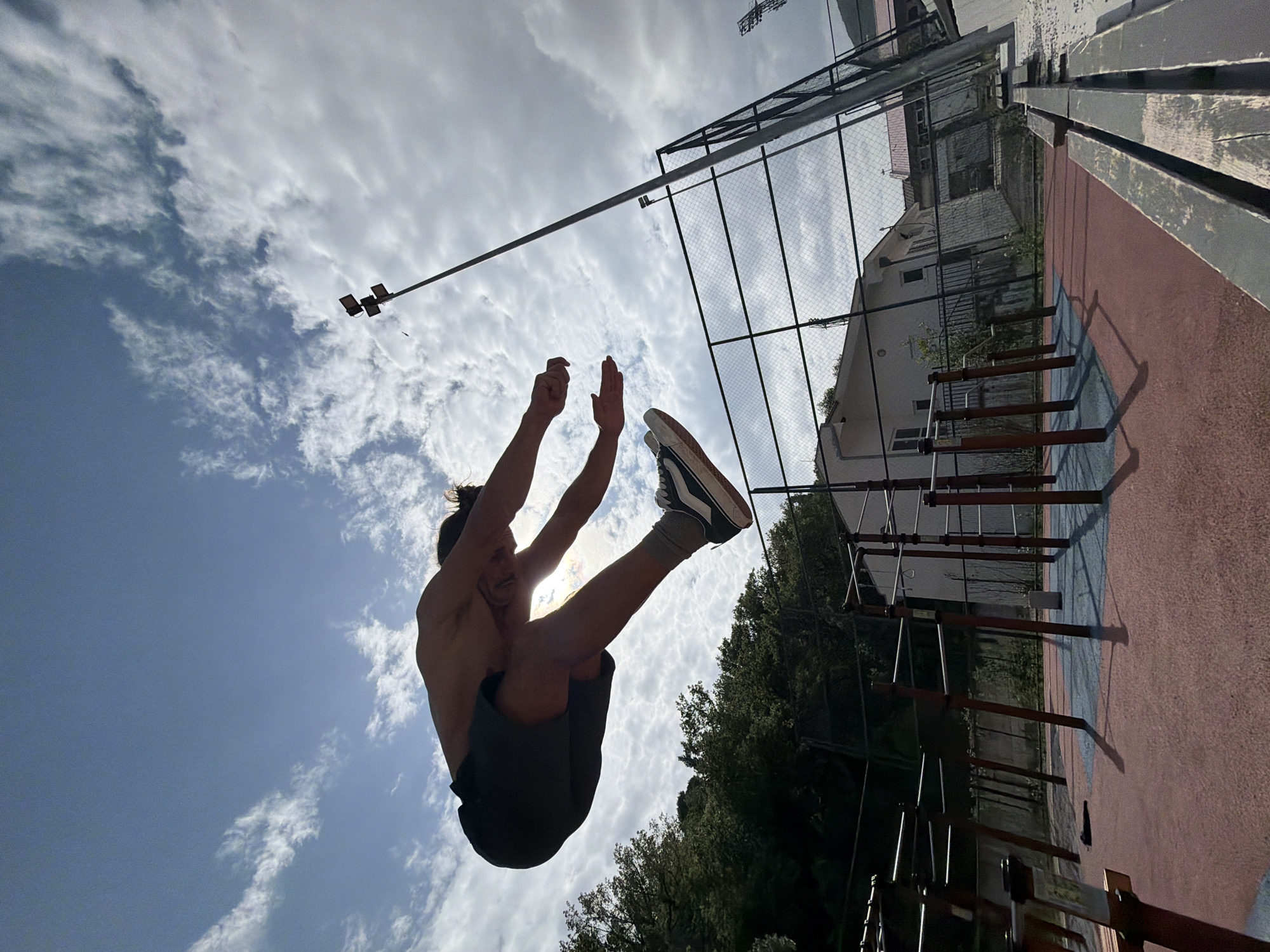 A man is captured mid-air in an acrobatic pose against a sky filled with clouds. He is wearing black shorts, gray socks, and black and white shoes. He is shirtless and his arms are extended upwards. In the background, there is a metal fence, a building with a white facade, green trees, and a red running track. The image appears to be taken at an outdoor fitness area in Petrovac na Moru, Montenegro.