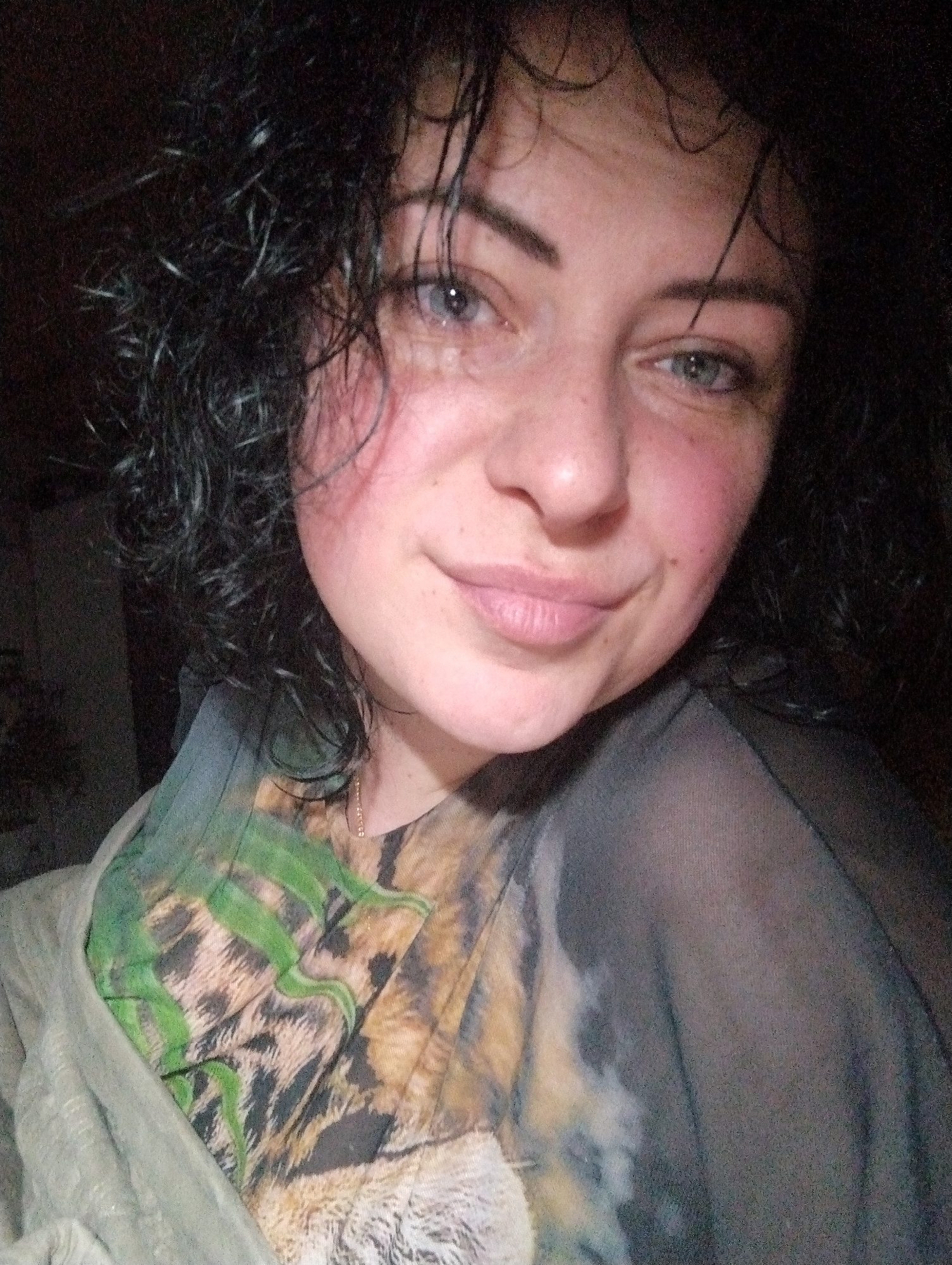 A person with wet, dark, curly hair and pale skin is facing the camera. The person's eyes are light-colored. They have a slight smile. The person is wearing a t-shirt with a tiger print and a light green garment draped over their shoulder. The background is dark. There is insufficient information to confirm the location in the city of Grishanki, Russia.