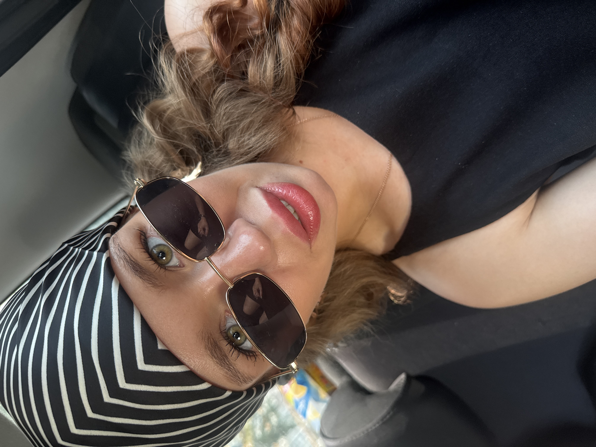 The image is a close-up, high-angle selfie of a young woman with light skin, green eyes, and reddish-brown curly hair. She is wearing a black headscarf with a bold white striped pattern, gold-rimmed sunglasses, and a black sleeveless top. Her lips are painted a glossy pink. A thin gold chain necklace is visible around her neck. The background appears to be the interior of a vehicle, with a dark grey seat on the left and a darker, possibly black, seat or surface on the right. The lighting suggests an outdoor environment, possibly during the day, as indicated by the natural light illuminating her face. There are no identifiable landmarks or elements that would place this image specifically in Chaikovsky, Russia.