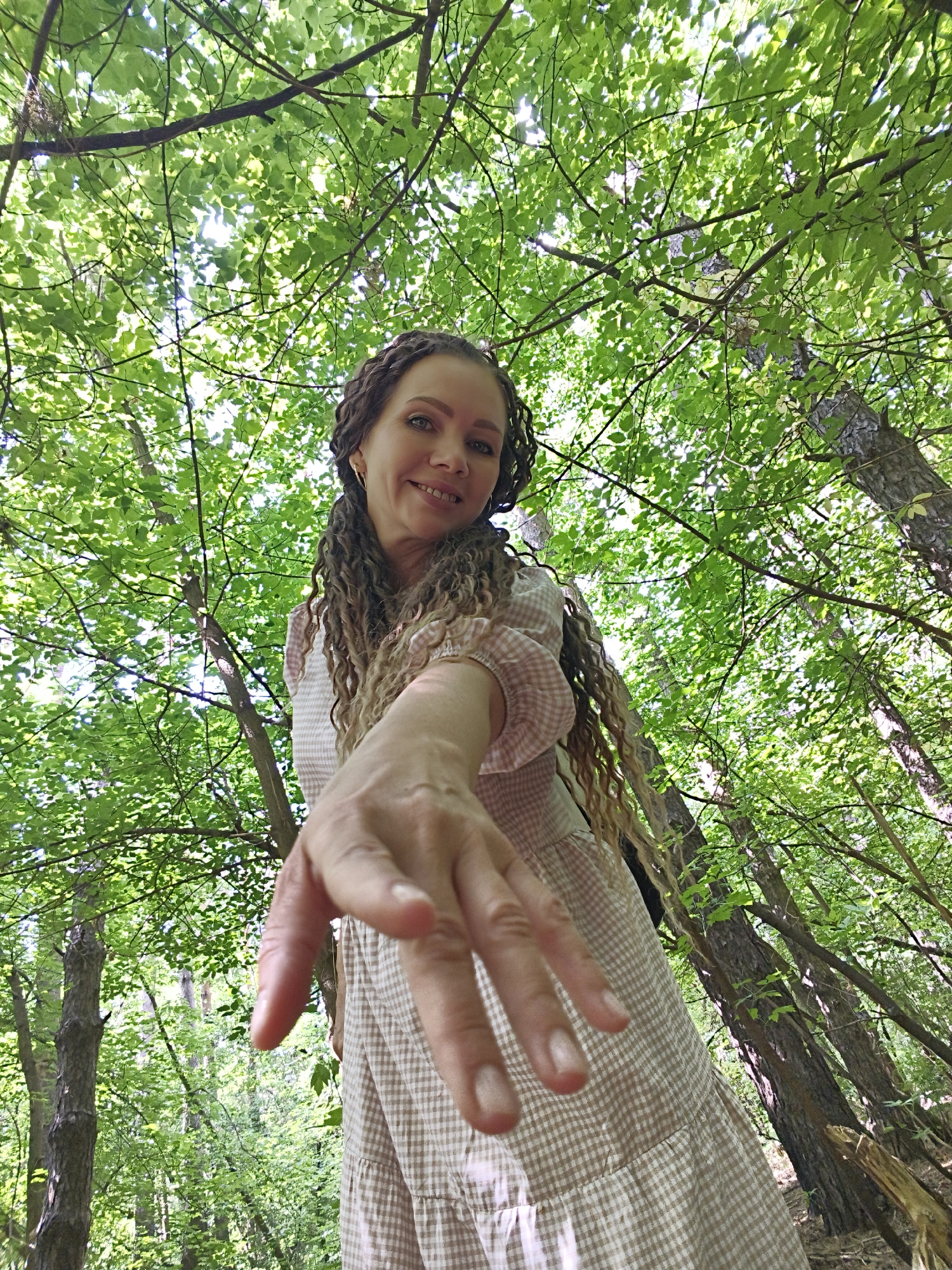 An overhead low-angle shot shows a woman with long, curly brown hair and fair skin wearing a light brown and white checkered dress. She is smiling at the camera with her head tilted slightly to her left and her right hand extended forward, palm facing the camera, fingers slightly curled. She is standing outdoors in a forest with lush green leaves and trees surrounding her, creating a dappled light effect. The woman is positioned in the lower right quadrant of the frame, while the upper two-thirds are dominated by foliage.