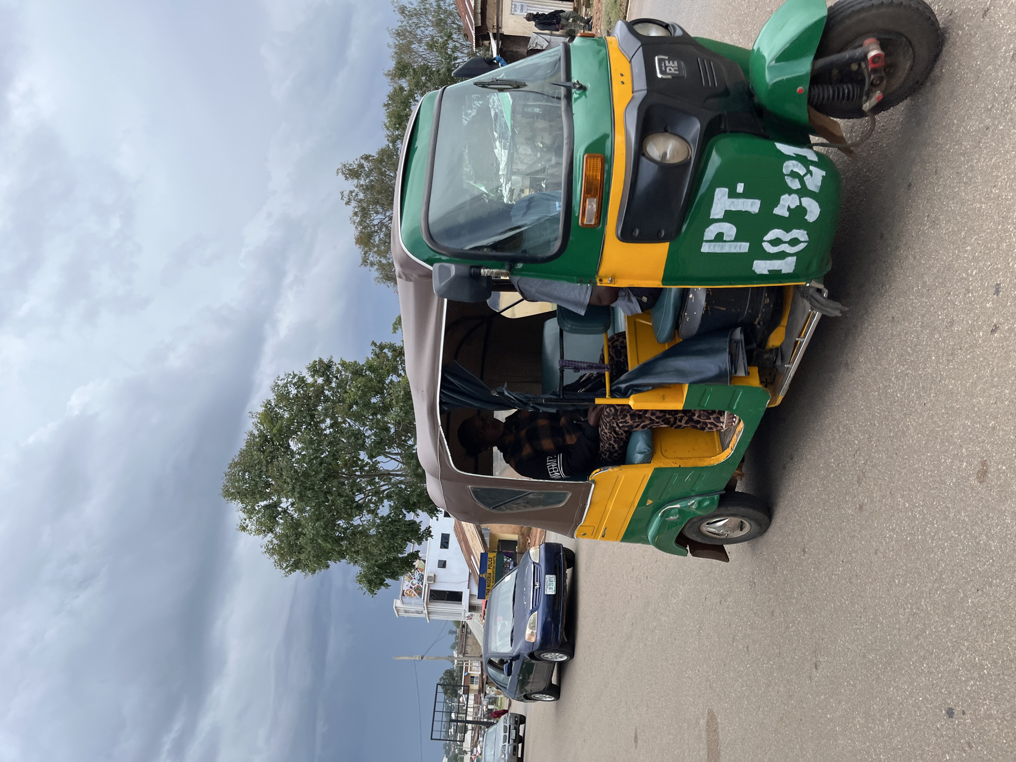 A green and yellow auto-rickshaw, identified with the registration "PT-18321," is parked on a paved road in Jos, Nigeria. A person wearing a plaid shirt and leopard-print pants is seated in the driver's area of the auto-rickshaw. In the background, a blue car is parked behind the auto-rickshaw, and further back, buildings are visible. One building has a sign that reads "THE NIGERIA POLICE." The sky is overcast, suggesting a cloudy day. The scene appears to be during daylight hours.