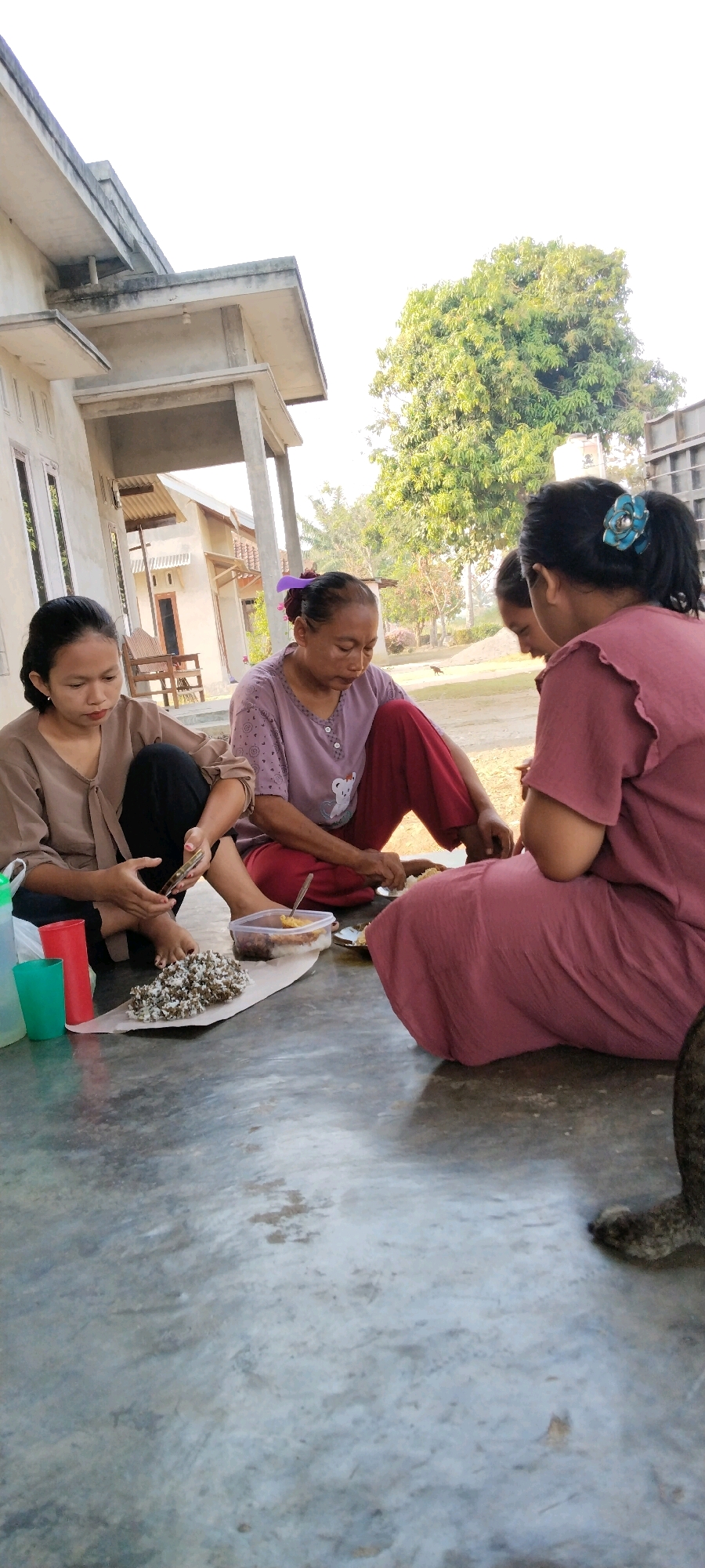 The image depicts three individuals, two women and one girl, seated on a concrete floor outside a building, possibly in Tanjung Ratu Ilir, Indonesia. The woman on the left is wearing a brown shirt and black pants, and she is looking down at her hands. The woman in the middle is wearing a purple shirt and red pants, and she appears to be eating. She has a purple hair tie in her hair. The girl on the right is wearing a pink dress and is partially obscured, facing away from the camera. In front of the group, on a sheet of paper, is a pile of rice and some food in a plastic container. Three plastic cups (green, red, and white) are also present. Behind the people, a building with a covered porch is visible, along with a large tree. The overall scene suggests a casual outdoor meal or gathering.