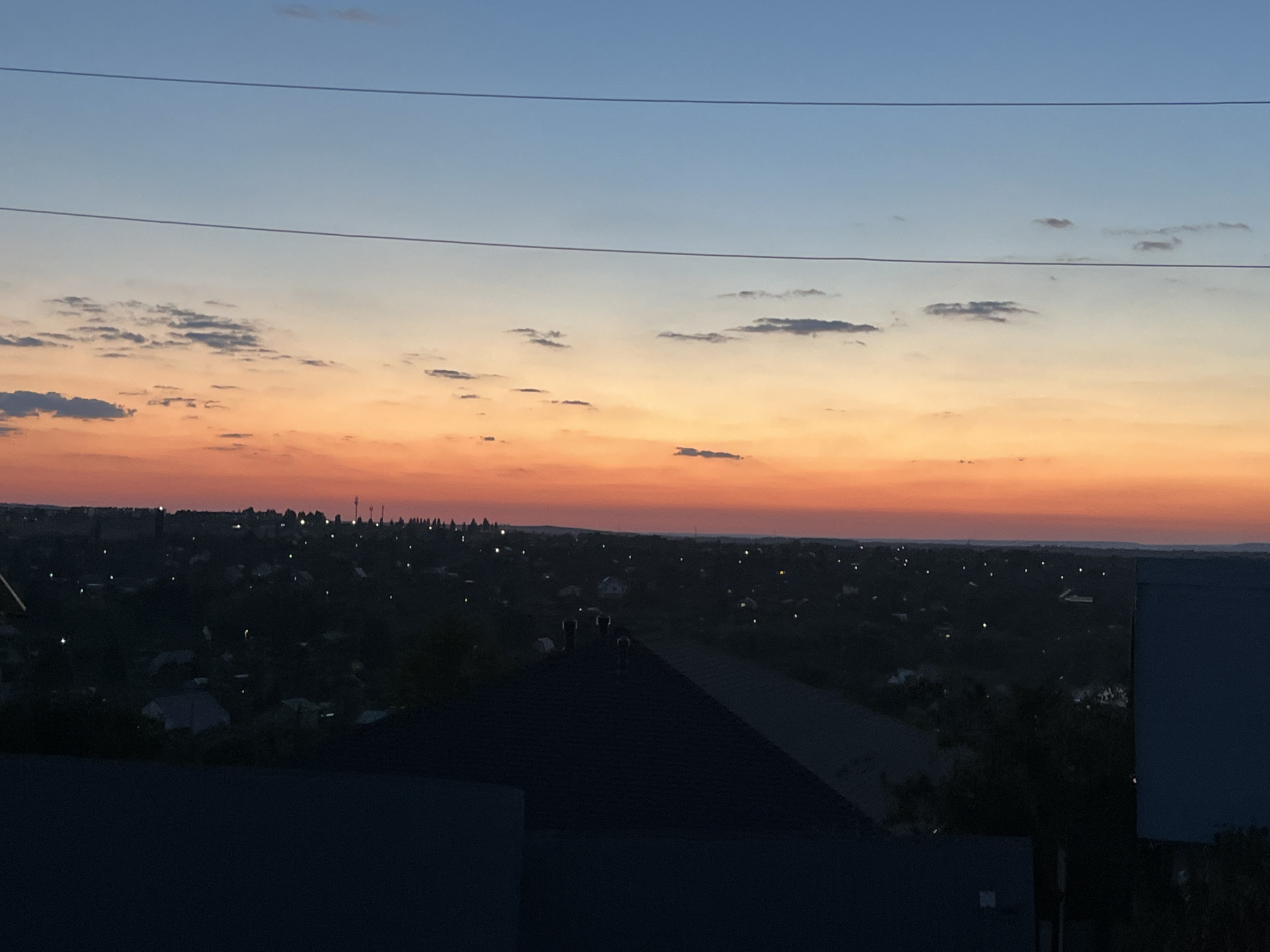 This is an outdoor, eye-level, medium shot of a sunset over a city. The sky is transitioning from a deep blue at the top to a vibrant orange and pink near the horizon, with scattered clouds. Two horizontal power lines stretch across the upper portion of the image. Below the sky, a dark silhouette of a city is visible, dotted with small lights. In the foreground, the dark shapes of rooftops and structures obscure the lower part of the landscape. The scene appears to be a residential area with houses and trees. The overall lighting suggests dusk.