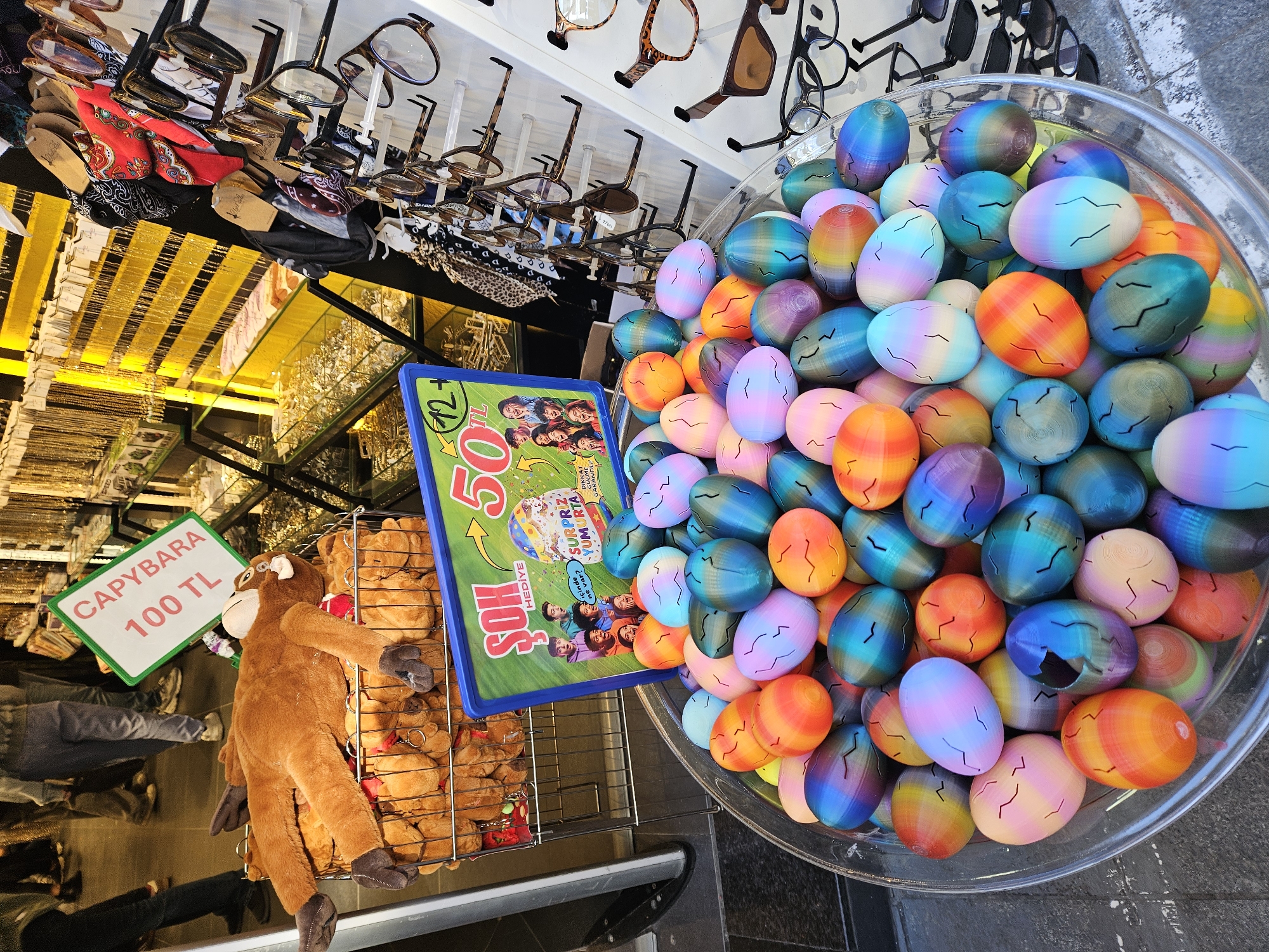 This image, taken in Kadıköy, Turkey, shows a vendor's stall with various items for sale. Overhead, numerous pairs of eyeglasses are displayed on stands. Below, a shelf holds gold-colored chains or necklaces.
To the left of the frame, a green sign indicates the price of a stuffed capybara at "100 TL". The plush toy itself hangs in front of a wire basket filled with more of the same capybara plushies.
Next to the capybara is a box with the text "SURPRIZ YUMURTA" and the number "50." To the right, a clear plastic bowl is overflowing with colorful, egg-shaped toys. These objects are arranged to attract the attention of potential customers. The setting appears to be a busy market or shopping area.