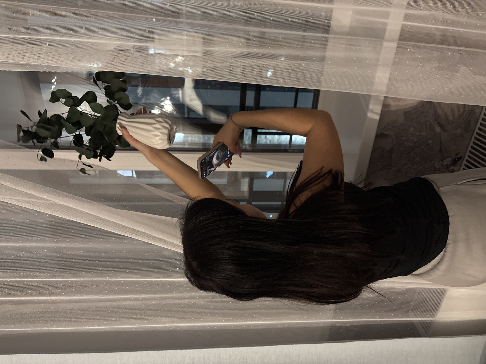 The image shows a woman indoors, standing near a window at what appears to be night or evening. She has long, dark hair and is wearing a black top and light-colored bottoms. She is captured from a rear, slightly elevated perspective.
With her right arm extended, the woman holds a white, vertically ribbed vase containing several green, leafy stems, resembling eucalyptus. Her left arm is also extended, holding a smartphone with a silver frame, which she is using to take a photograph of the plant arrangement. The screen of the phone is visible but too small to discern any specific content or text.
Behind the woman and the plant, sheer white curtains with a subtle dotted pattern are drawn across a window. Through the semi-transparent curtains and the windowpane, the outside environment appears dark, with indistinct reflections of interior lights or distant exterior light sources. Below the window, a part of a wall or window sill and what looks like a heating vent or radiator are visible.
The scene suggests a moment of indoor activity, possibly in a home or apartment in Yekaterinburg, Russia, where the woman is staging and photographing a decorative plant. No explicit text is visible in the image.