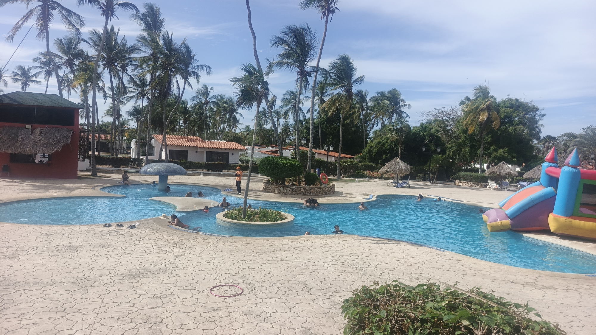 The image shows a swimming pool complex at Playa El Agua, Venezuela. The complex consists of several interconnected pools filled with blue water. People are seen swimming and relaxing in the pools. On the right, an inflatable water slide is visible next to the pool. Buildings with terracotta roofs and palm trees are in the background. The pool deck is made of textured concrete. In the foreground, there is a green bush. The sky is partially cloudy.