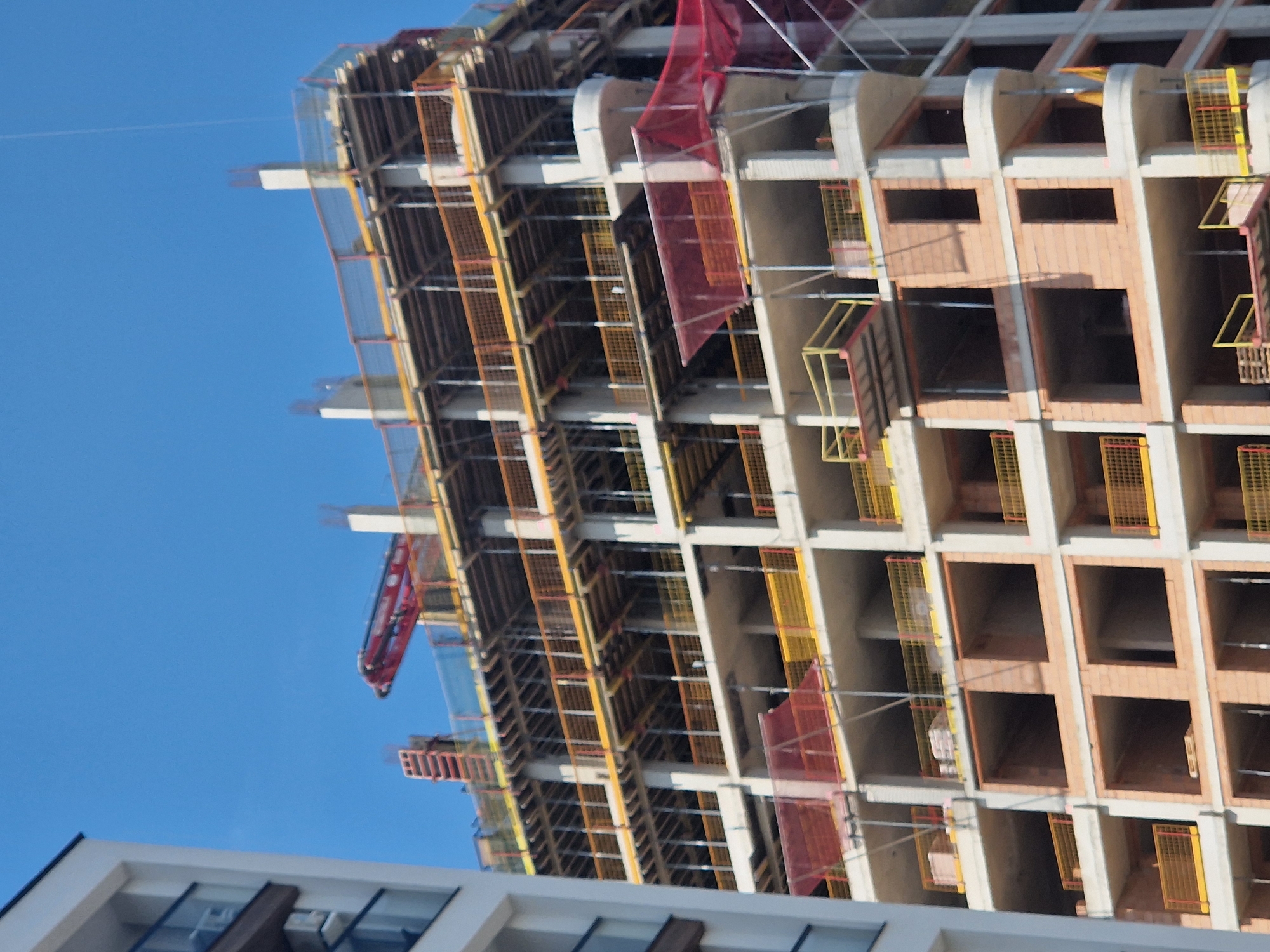 The image shows a building under construction in the city of Unknown, Unknown. The building is a high-rise with a concrete frame and partially bricked-in window openings. The construction site is secured with yellow and orange mesh safety netting. A red construction crane is visible on the side of the building. The sky is blue and clear. In the foreground, a portion of a white, multi-story building with dark windows can be seen.