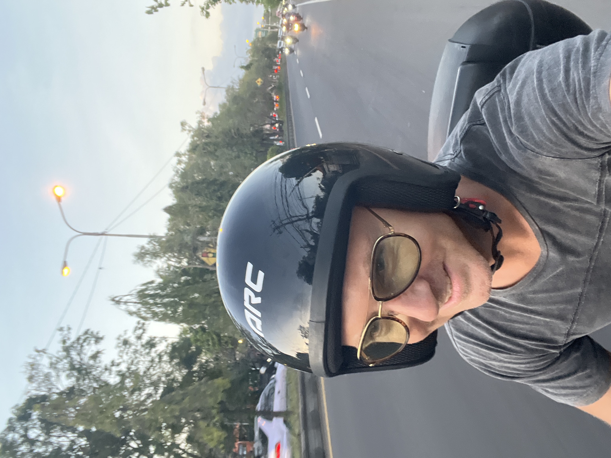 This media file is a selfie taken from the perspective of someone riding a motorcycle. The subject is wearing a black helmet with the white text "ARC" visible on the side, and sunglasses with gold frames. They are also wearing a grey t-shirt. The scene is a road with other motorcycles and cars visible in the background, suggesting a busy street. Trees line the side of the road, and streetlights are present, one of which is illuminated. The sky appears overcast, indicating it might be dusk or a cloudy day. The overall impression is of someone traveling on a motorcycle in an urban environment, possibly in Denpasar, Indonesia, given the location context. The selfie-style composition and the subject's relaxed posture suggest a casual and possibly adventurous mood.