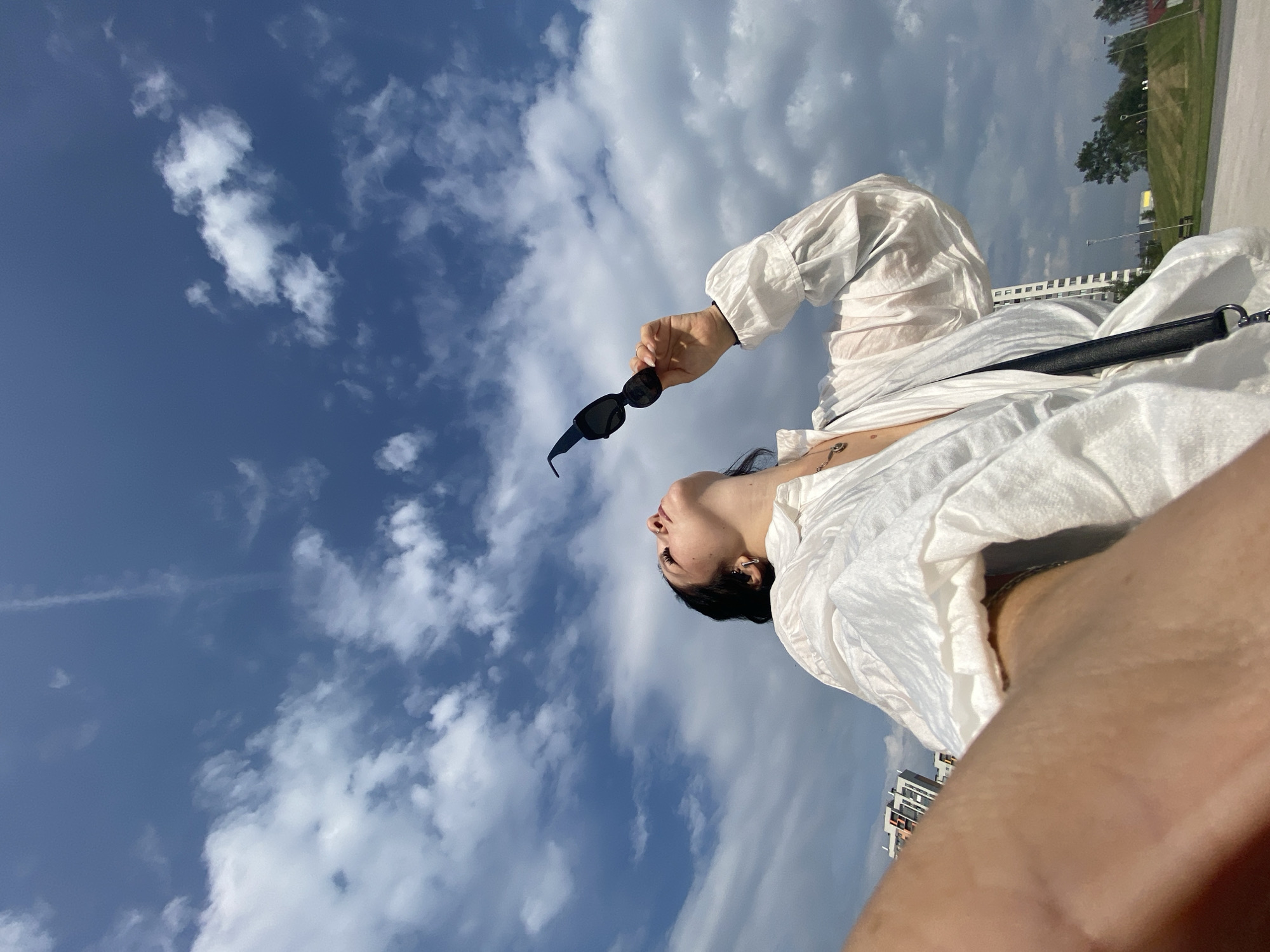 A low-angle, selfie-style shot shows a woman looking up at a cloudy blue sky. She is wearing a white, loose-fitting shirt with puffed sleeves and a delicate necklace. In her right hand, she holds a pair of black sunglasses up towards the sky. Her left arm is out of frame, but a portion of her skin is visible in the foreground. In the background, there are several tall buildings, some trees, and a grassy hill. The overall setting appears to be an outdoor urban environment.