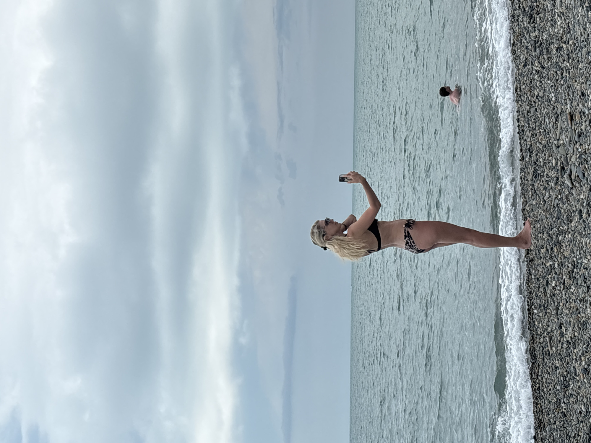 This media file captures a vertical, full-body shot of a woman in a bikini standing on a pebble beach and taking a selfie with her phone. The woman has blonde hair and is wearing sunglasses. She is positioned in the mid-ground, facing away from the viewer, with her right arm extended holding the phone. In the background, the ocean stretches out to the horizon under a cloudy sky. A man is visible in the water, further out from the shore. The scene suggests a casual day at the beach, likely during daylight hours given the lighting. The weather appears overcast, contributing to a soft, diffused light. The pebble beach is a prominent feature, with the pebbles appearing dark and rounded. The ocean has gentle waves rolling in, creating a white foamy edge where it meets the shore. The overall atmosphere is relaxed and summery. No visible text is present in the image.