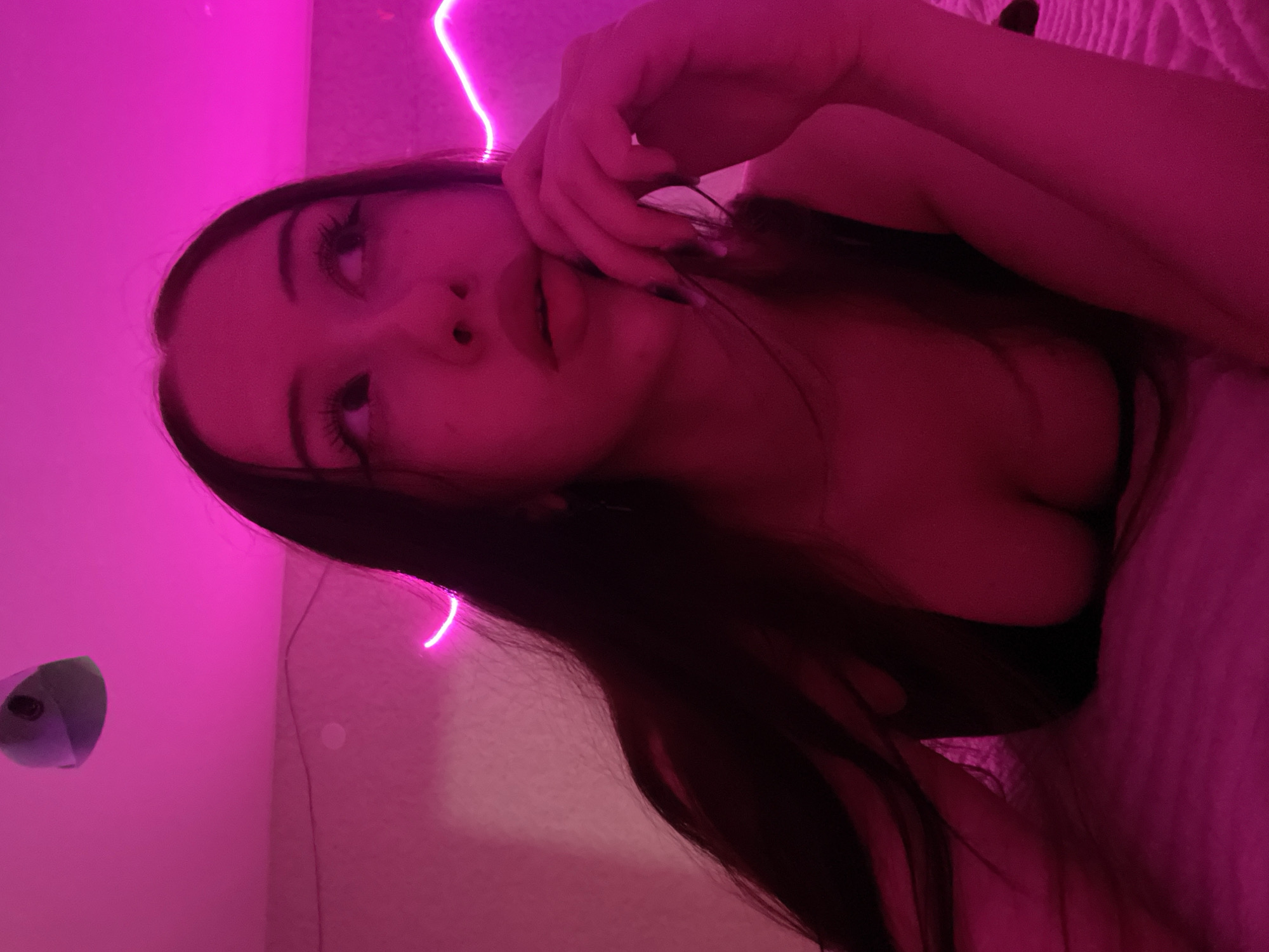 The image shows a young woman lying down in a room illuminated by pink neon lights. She has long, dark hair and is wearing a dark top. Her hand is near her mouth, and she is looking upwards and to the side. The background shows a wall with some sort of light fixture or decoration. There is no discernible information within the image to determine the location, including the city of Votkinsk, Russia.