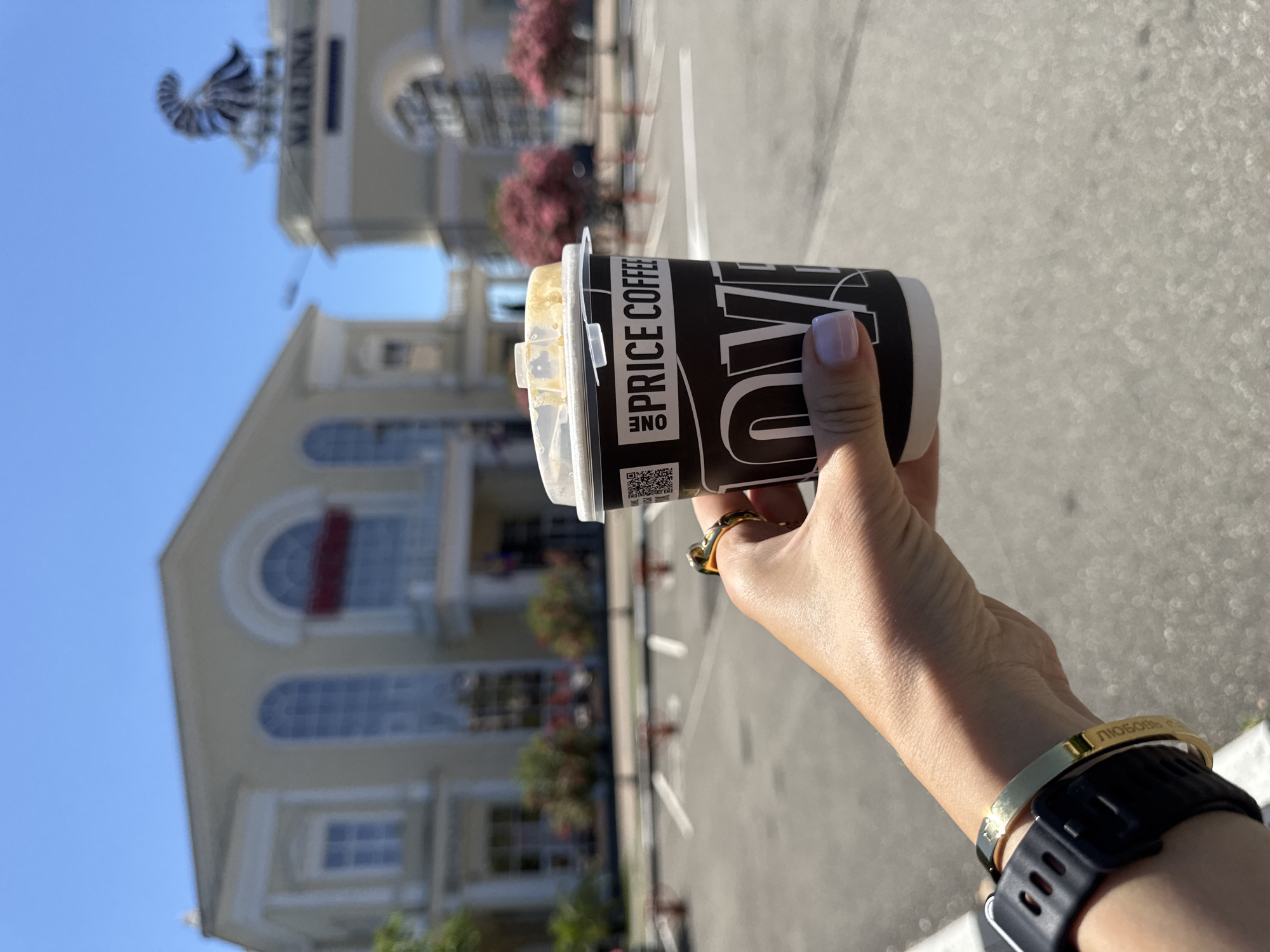 A first-person perspective shows a hand holding a black paper coffee cup with white lettering that reads "ONE PRICE COFFEE" and "LOVE". The cup has a white lid with some dried coffee residue. The hand is adorned with a gold ring on the ring finger and a gold bracelet with engraved Cyrillic text that appears to read "ЛЮБОвь" (Lyubov, meaning love) and a black watch on the wrist.
In the background, a light-colored, neoclassical building with arched windows and columns is visible under a clear blue sky. To the right, another arched structure, possibly an entrance, bears the word "MARINA" in blue lettering and is topped with a decorative, fan-shaped sculpture. Pink flowering trees are scattered throughout the scene, and a parking lot with white markings is present in the foreground. This scene is consistent with the entrance area of the "Morskoy Voyennyy Gospital" (Naval Military Hospital) in Sochi, Russia, which is often referred to as the "Marina" due to its proximity to the sea and its naval association.