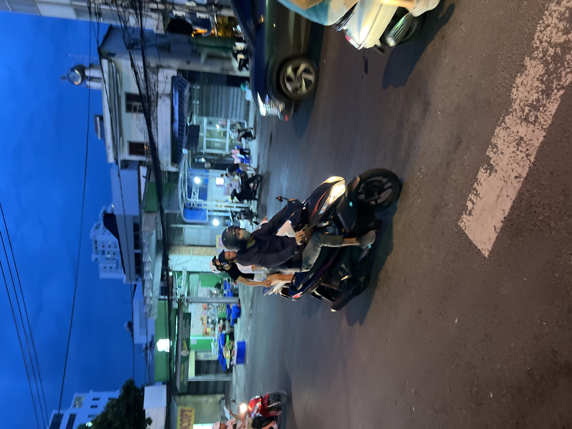 In the city of Nha Trang, Vietnam, at dusk, several people are riding motorcycles on a street. The sky is a deep blue, and streetlights are illuminated.
In the foreground, a person wearing a dark jacket and a helmet is riding a dark-colored motorcycle. Another person, also wearing a helmet, is riding pillion behind them. A third person, wearing a white helmet and a dark shirt, is riding pillion behind the second person. The motorcycle's headlights are on.
To the left, a red motorcycle with two occupants, both wearing helmets, is moving away from the viewer. The person in front is wearing a white shirt and a helmet, and the person behind is wearing a white shirt and a helmet.
In the background, a dark-colored car is visible, as is a white scooter with its rear visible. Buildings line the street, some appearing to be businesses with awnings and lights. A few people are visible standing or sitting near the businesses. The street surface is asphalt, with white markings indicating a pedestrian crossing. Power lines are strung overhead. The overall scene suggests a busy street in a Vietnamese city at twilight.