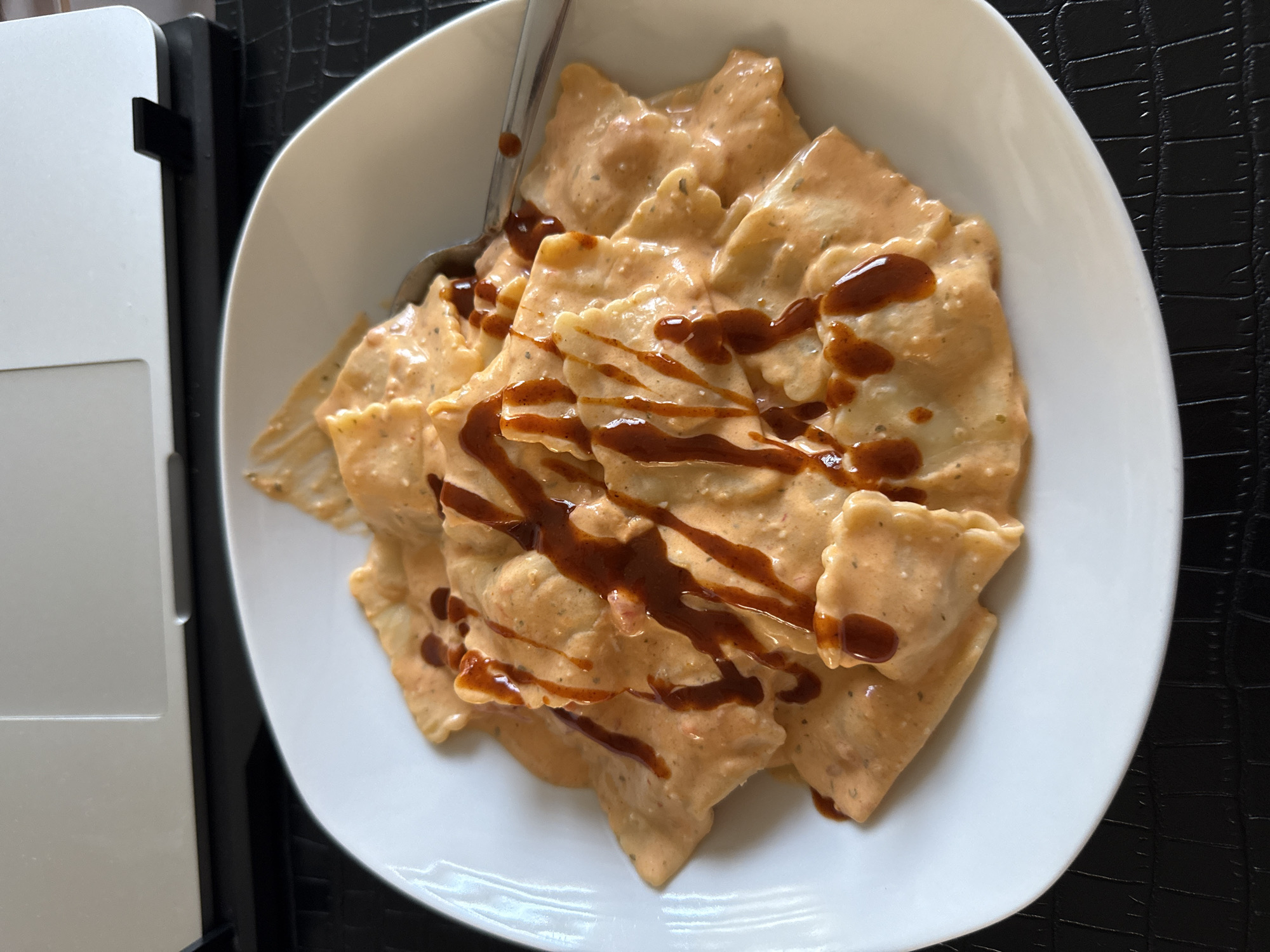 In Marum, Netherlands, a close-up, overhead shot captures a plate of ravioli. The ravioli are coated in a creamy, orange sauce and drizzled with a darker sauce, possibly barbecue sauce. A silver spoon rests on the right side of the plate, partially submerged in the pasta. The plate sits on a black, textured surface, which appears to be a placemat or tabletop. In the background, the edge of a silver laptop is visible, positioned on a black surface. The lighting suggests an indoor environment, possibly a home or office setting, during daylight hours. There are no people visible in the frame.