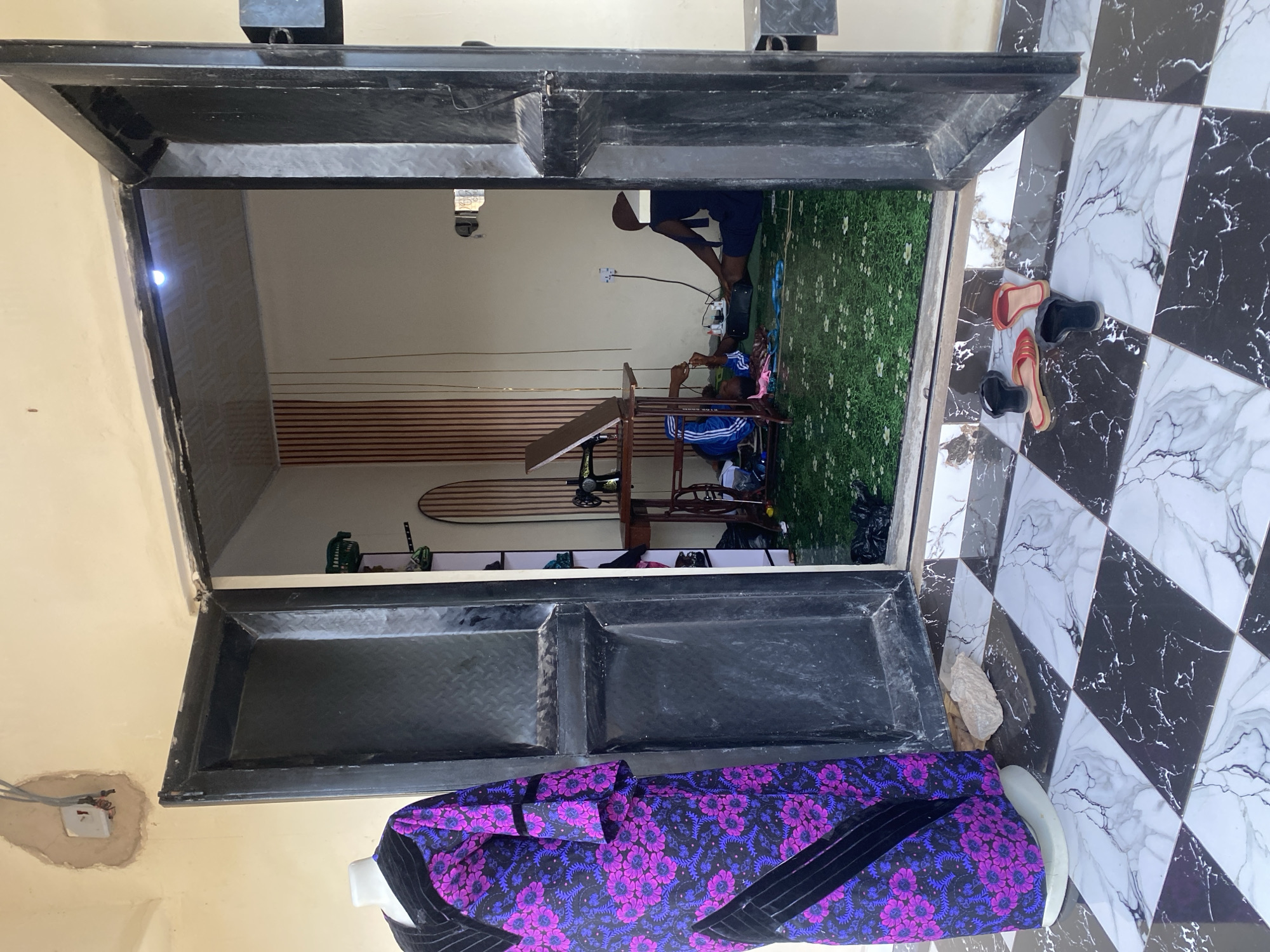 In Jauro Yinu, Nigeria, a doorway opens into a modest workshop. On the left, a dress form displays a long, patterned garment with purple floral designs and black trim. The doorway itself is framed by thick, dark metal doors, slightly ajar. The floor inside is covered in a green carpet with a white floral pattern. Within the workshop, a sewing machine rests on a wooden table. A person, partially visible and wearing a dark head covering and blue shorts, sits on the floor, appearing to be working on something with their hands. Another individual, in blue and white striped athletic pants, is also on the floor, with their legs bent. Various items, including a black bag, a white bottle, and spools of thread, are scattered around them. In the background, a wall is decorated with vertical wooden slats, and a light fixture is visible on the ceiling. Outside the workshop, on the black and white checkered tiled floor, a pair of red sandals and two black flip-flops lie near the entrance.
