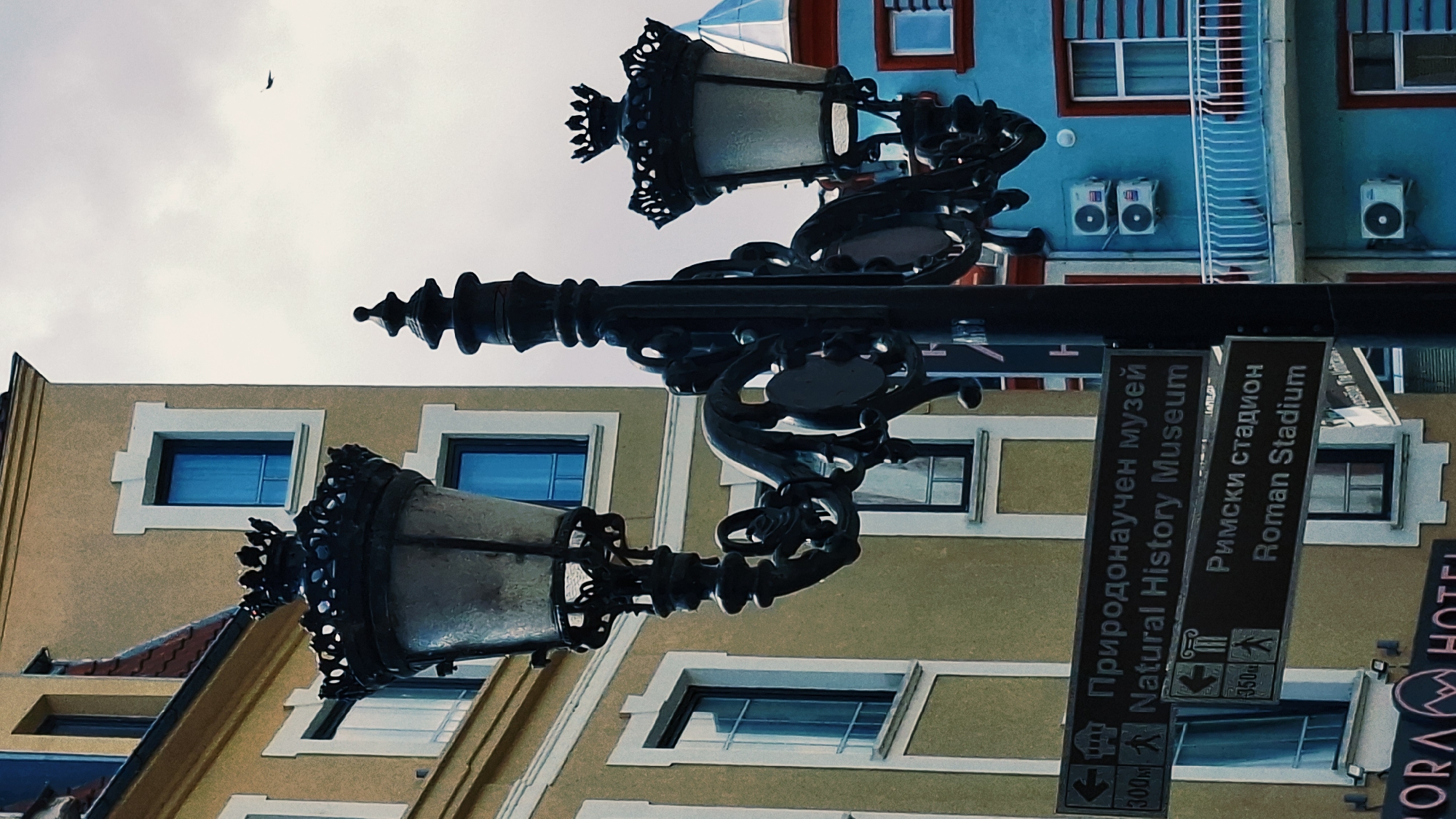 This is a close-up, upside-down photograph taken outdoors in Plovdiv, Bulgaria, focusing on two ornate street lamps and a directional signpost. The street lamps are made of dark metal, featuring decorative scrollwork and a textured glass shade, suggesting an older, possibly historical, part of the city.
The signpost displays directions in both Bulgarian and English. One sign points to "Природонаучен музей Natural History Museum" with an arrow indicating 300 meters. Another sign points to "Римски стадион Roman Stadium" with an indication of 350 meters. The presence of these signs strongly suggests a tourist-oriented area within the city.
The buildings in the background appear to be in different architectural styles and colors. One building on the left has a beige facade with white-trimmed windows, while a building on the right is painted a vibrant blue with red trim around the windows, and features external air conditioning units and a ladder. The sky above is overcast, indicating a cloudy day, and a small bird is visible in flight. The composition is dominated by the street lamps and the signpost, creating a sense of urban exploration and highlighting key points of interest within the city. The overall mood is one of discovery and a hint of historical charm.