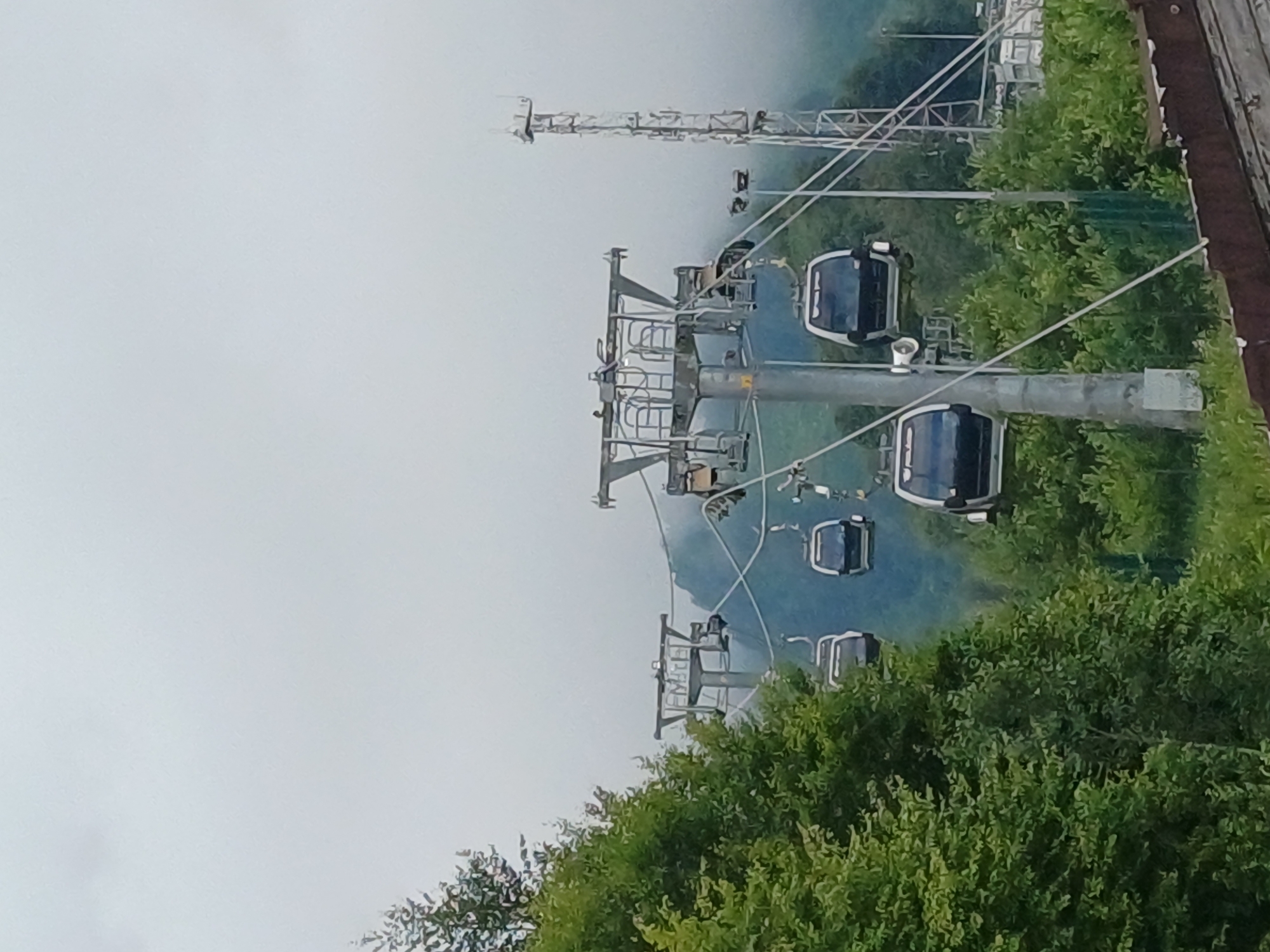 The image shows a cable car system operating in Estosadok, Russia. Several cable car cabins are visible, suspended from wires and moving between pylons. The cable cars are modern, enclosed cabins with windows. The pylons are tall, metallic structures supporting the cables. Lush green trees and vegetation surround the cable car infrastructure, indicating a mountainous or hilly terrain. In the background, a tall, slender tower, possibly a communication tower or a part of the cable car system, is visible. The sky is overcast and hazy, suggesting cloudy weather. The overall scene depicts an outdoor recreational or transportation facility in a natural environment.