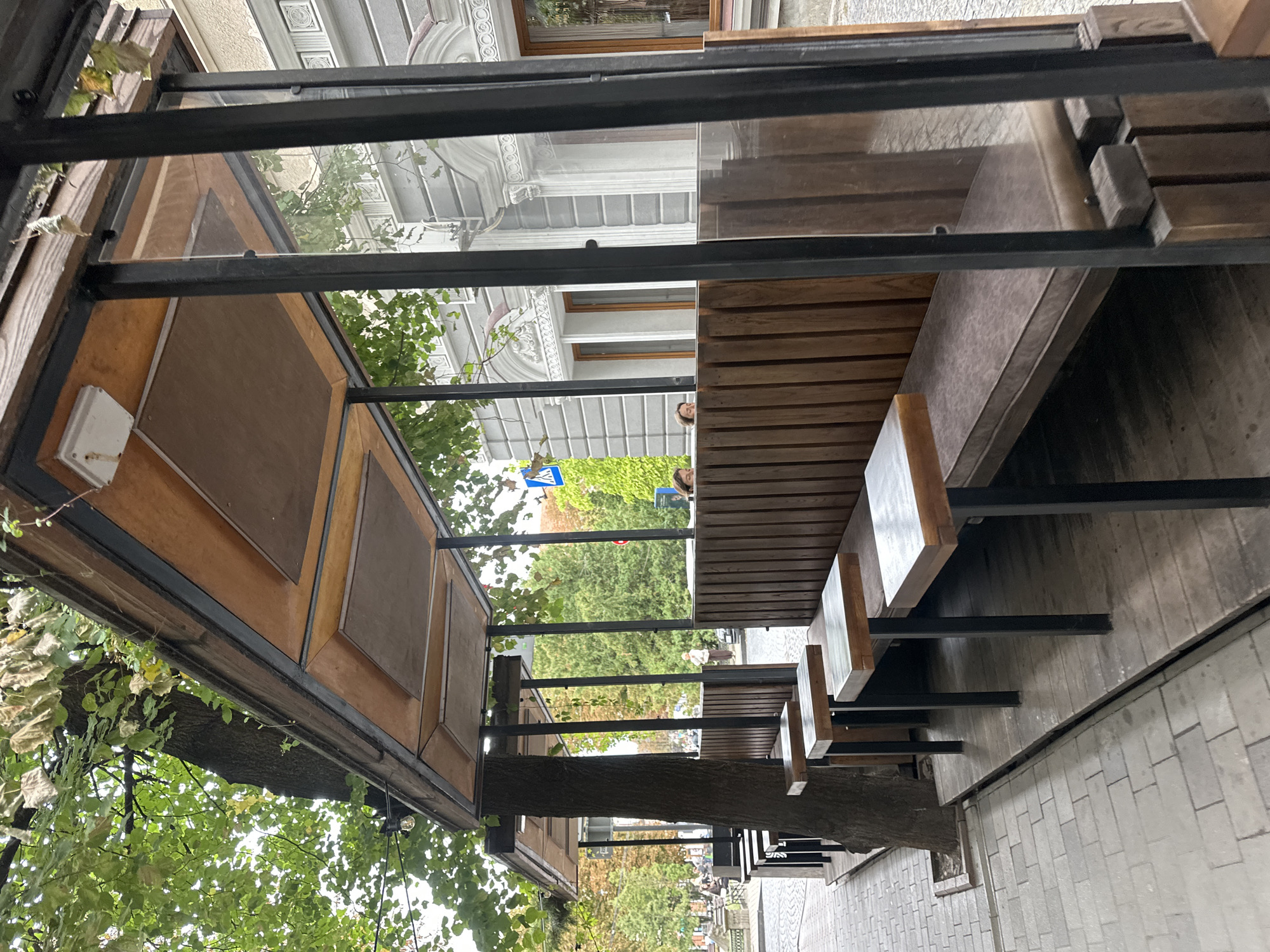 This image depicts an outdoor seating area for a cafe or restaurant, located in Ternopil, Ukraine. The seating area features a wooden structure with a sloped roof and glass panels on the sides. Inside, there are long wooden benches with cushions and small wooden tables supported by black metal legs. A large tree trunk is integrated into the structure, with its branches extending overhead and leafy green foliage partially obscuring the roof. The ground beneath the seating area is wooden, and the surrounding walkway is paved with grey bricks. In the background, a building with ornate architectural details and windows can be seen. Two individuals are partially visible inside the seating area, suggesting it is operational. The overall scene conveys a relaxed, urban outdoor dining atmosphere.