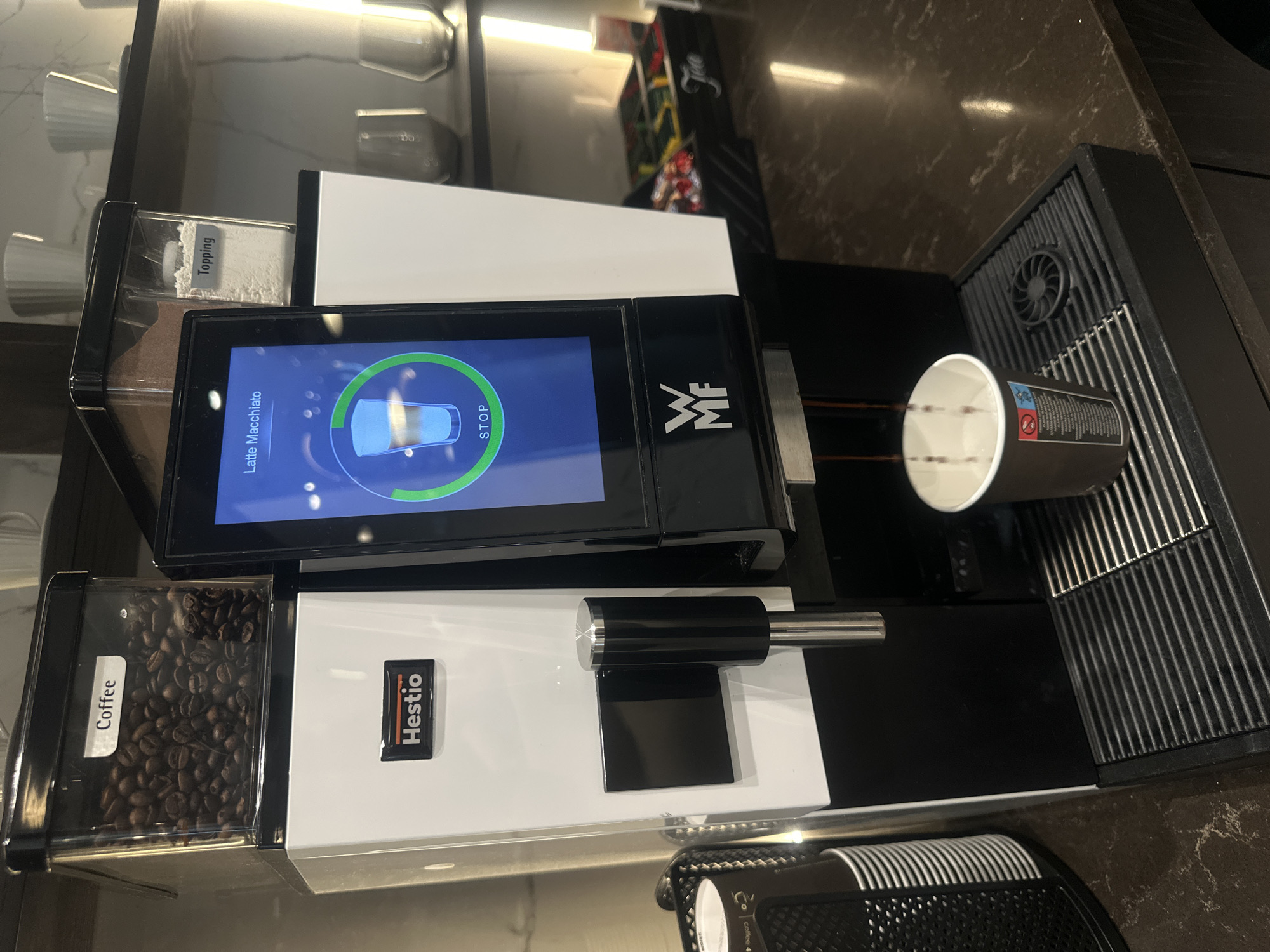 The image shows a WMF coffee machine preparing a latte macchiato. The machine has containers for coffee beans and topping. A cup is positioned on the drip tray. The machine's display indicates the latte macchiato is being prepared. The brand "Hestio" is visible on the machine. The scene appears to be indoors, likely in a cafe or office break room. There are no people visible. The location is unspecified but based on available information is in Riga, Latvia.