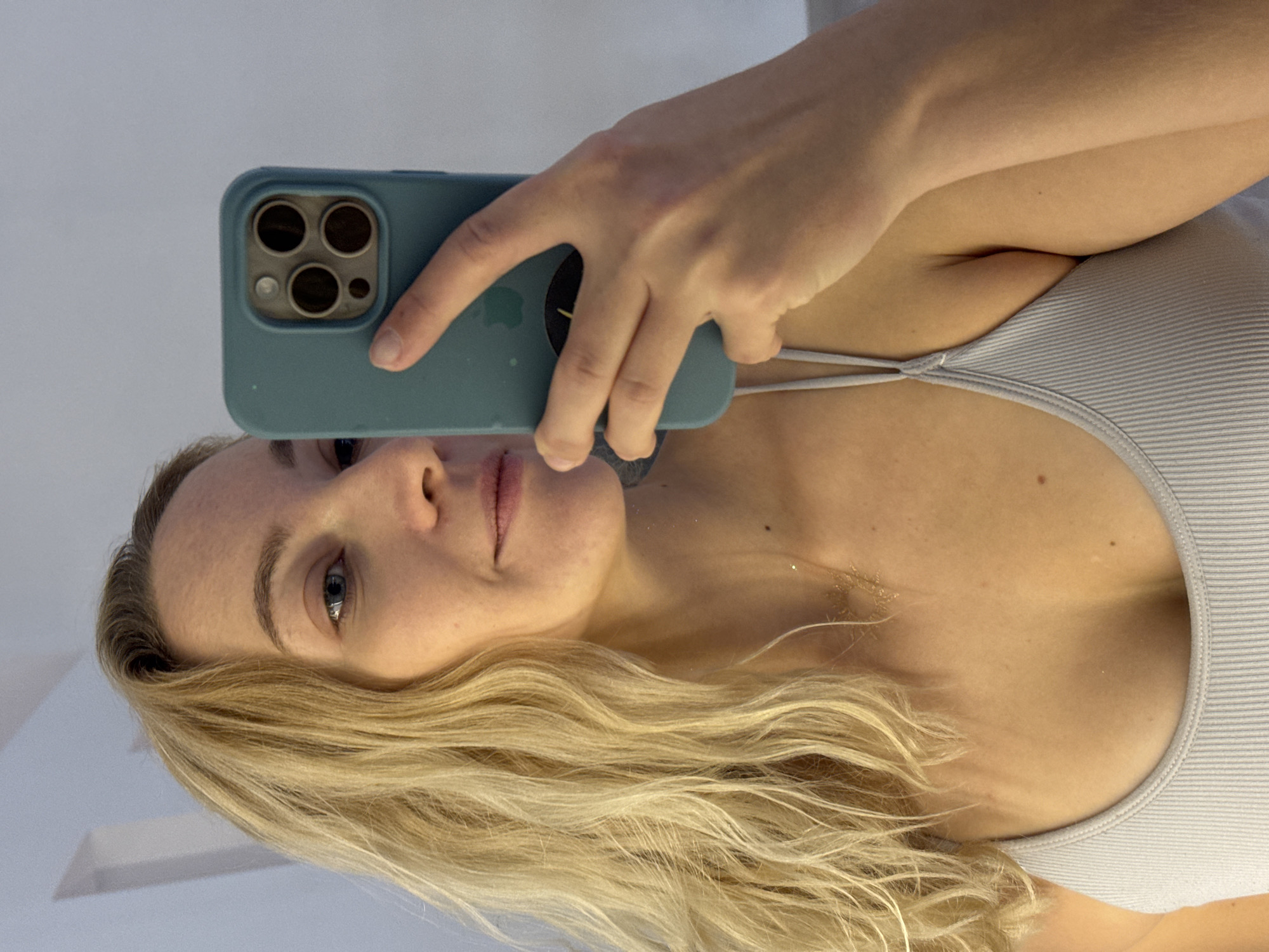 This is a medium-angle, indoor, selfie-style photograph of a fair-skinned woman with blonde, wavy hair, looking into a mirror. She is wearing a light-colored, ribbed tank top. She is holding a light blue smartphone in her right hand, partially obscuring her face. The background is a plain, lightly colored wall. There is no discernible information about the location in Minsk, Belarus.
