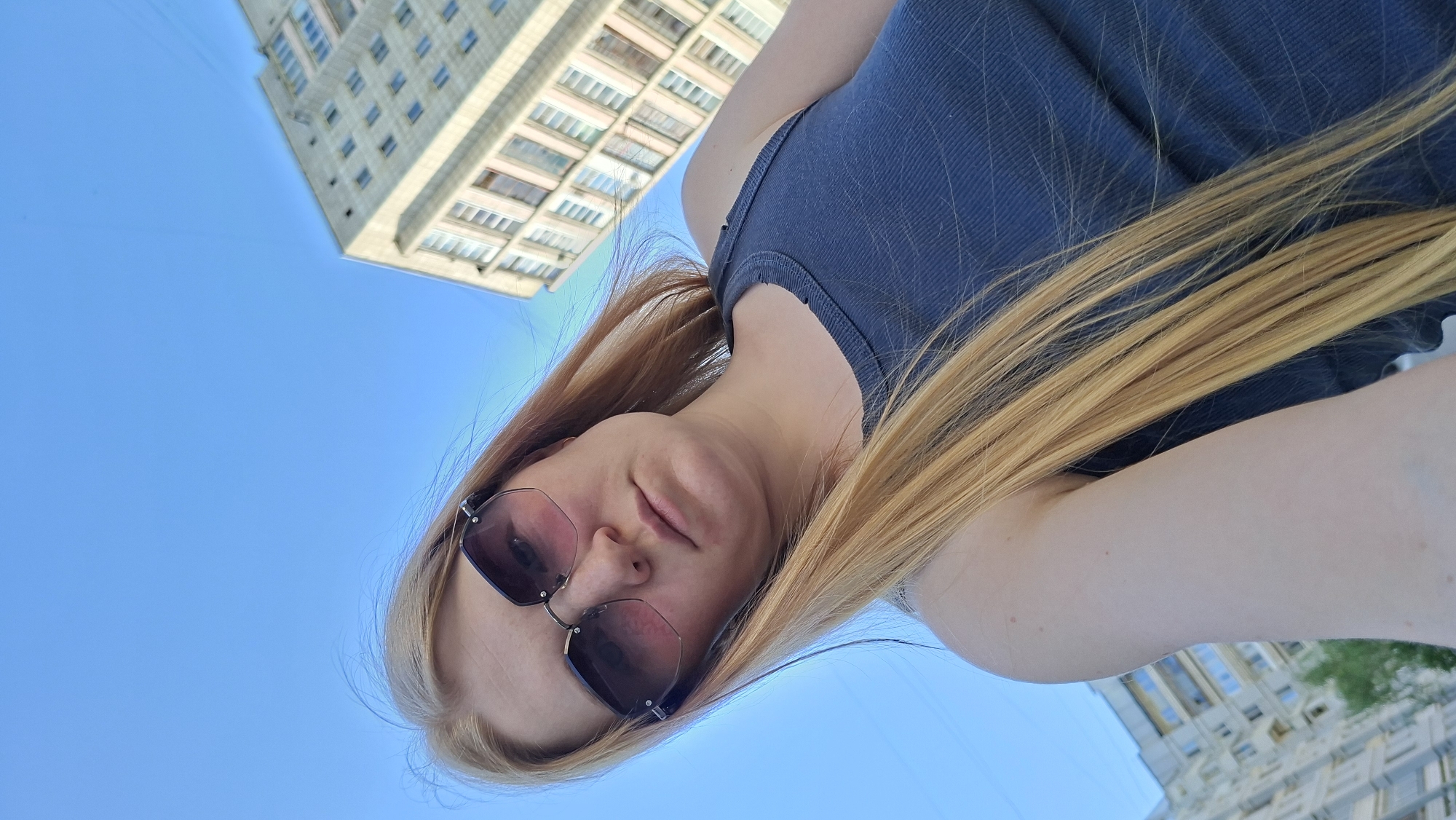 A low-angle, rotated selfie shows a fair-skinned person with long, light blonde hair wearing dark-framed sunglasses. The person is wearing a blue tank top. The background reveals a blue sky and two buildings. The building in the upper left has multiple windows, while the building in the lower right is partially visible. The location is Chaikovsky, Russia.