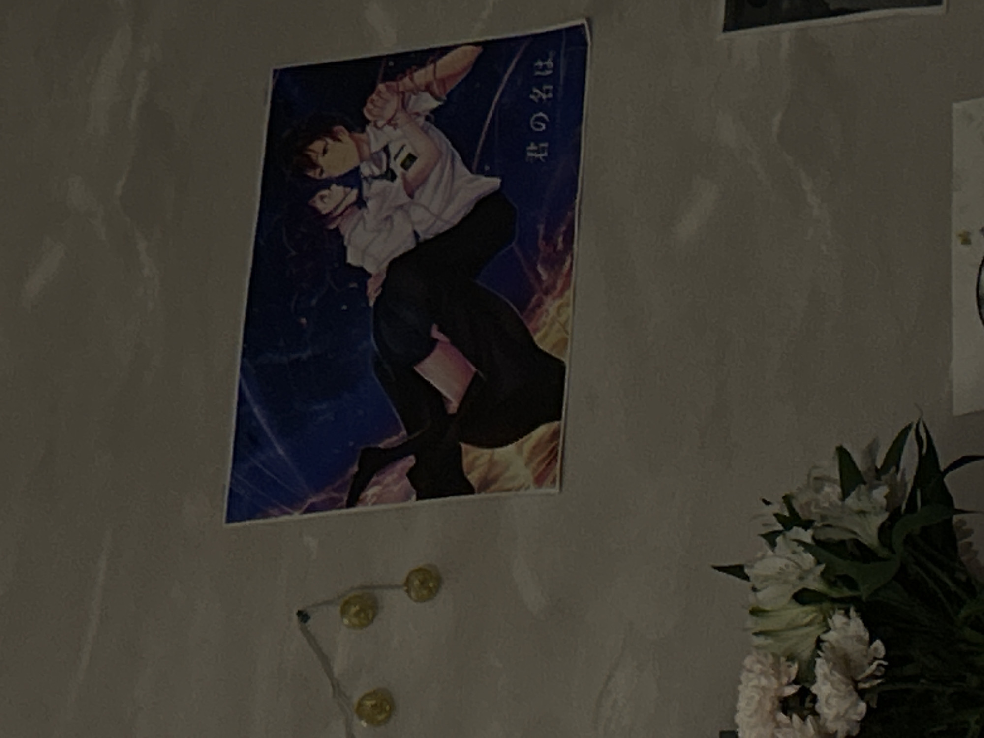 This image, taken in Chaikovsky, Russia, displays an indoor scene featuring a poster, string lights, and a bouquet of flowers against a wall.
The central subject is a vertical poster affixed to a light-colored, possibly textured, wall. The poster features anime-style artwork, depicting two figures in a dynamic pose. One figure, appearing to be a male, wears a white collared shirt and dark trousers, holding or embracing a female figure. The background of the poster is a dark, dramatic sky with celestial elements and an orange glow near the bottom. Vertically aligned Japanese text "君の名は" (Kimi no Na wa), which translates to "Your Name," is visible on the upper right side of the poster.
Below the poster, a string of small, round, yellow lights, resembling fairy lights, is draped along the wall. To the bottom right of the frame, a bouquet of fresh flowers, predominantly white with green foliage, is partially visible. The flowers appear to be lilies or a similar variety.
The lighting in the scene is dim and atmospheric, suggesting it is evening or nighttime, with artificial light sources creating soft reflections and shadows on the wall. The overall impression is that of a personalized and cozy indoor space.