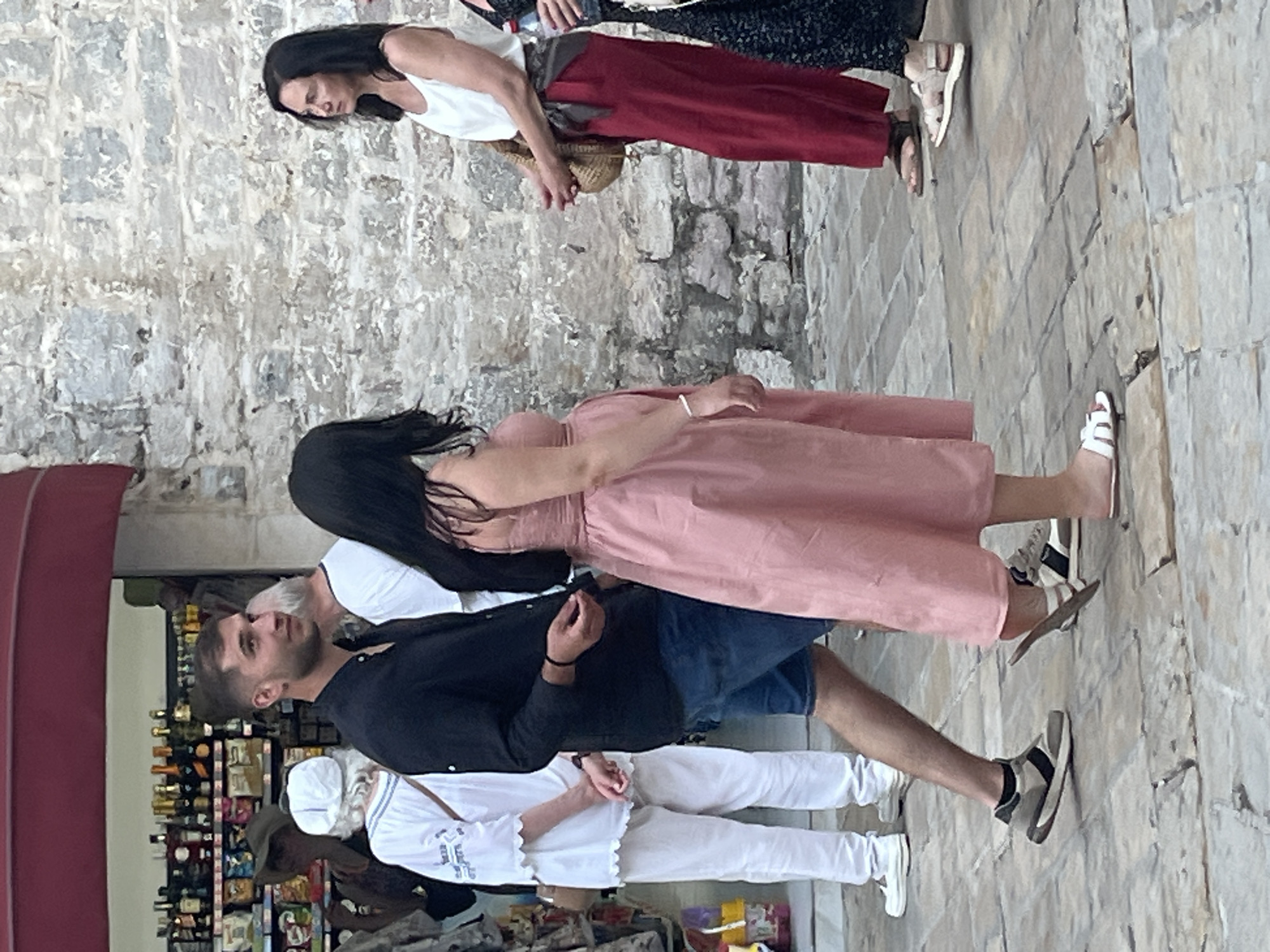 The image captures a street scene in Budva, Montenegro, focusing on a group of people interacting near a building with a stone facade. In the foreground, a man in a black shirt and denim shorts is walking, glancing to his left. To his right, a woman in a pink dress is walking in the same direction, also looking to her left. Behind them, a woman in a white tank top and red trousers stands facing right, holding a straw bag. To the far right, a portion of another woman in a black patterned dress is visible. In the background, a shop display filled with bottles and packaged goods is visible under a red awning. The ground is paved with stone slabs.