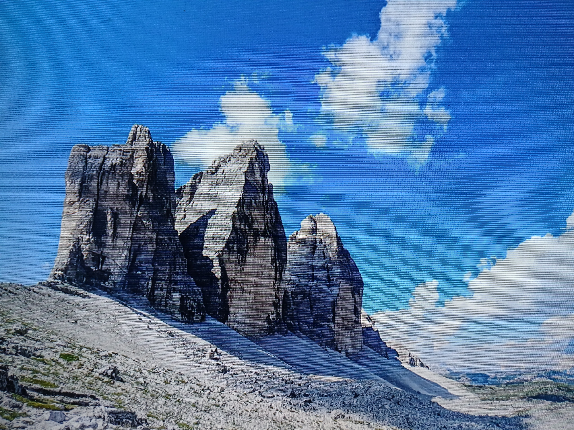 The image depicts a mountain range with three distinct peaks under a partly cloudy blue sky. The peaks appear rocky and striated, with shades of gray and brown. The terrain leading to the base of the peaks is rocky and sloped, sparsely covered with vegetation. There are no people visible in the image. The location is not Juan Griego, Venezuela.