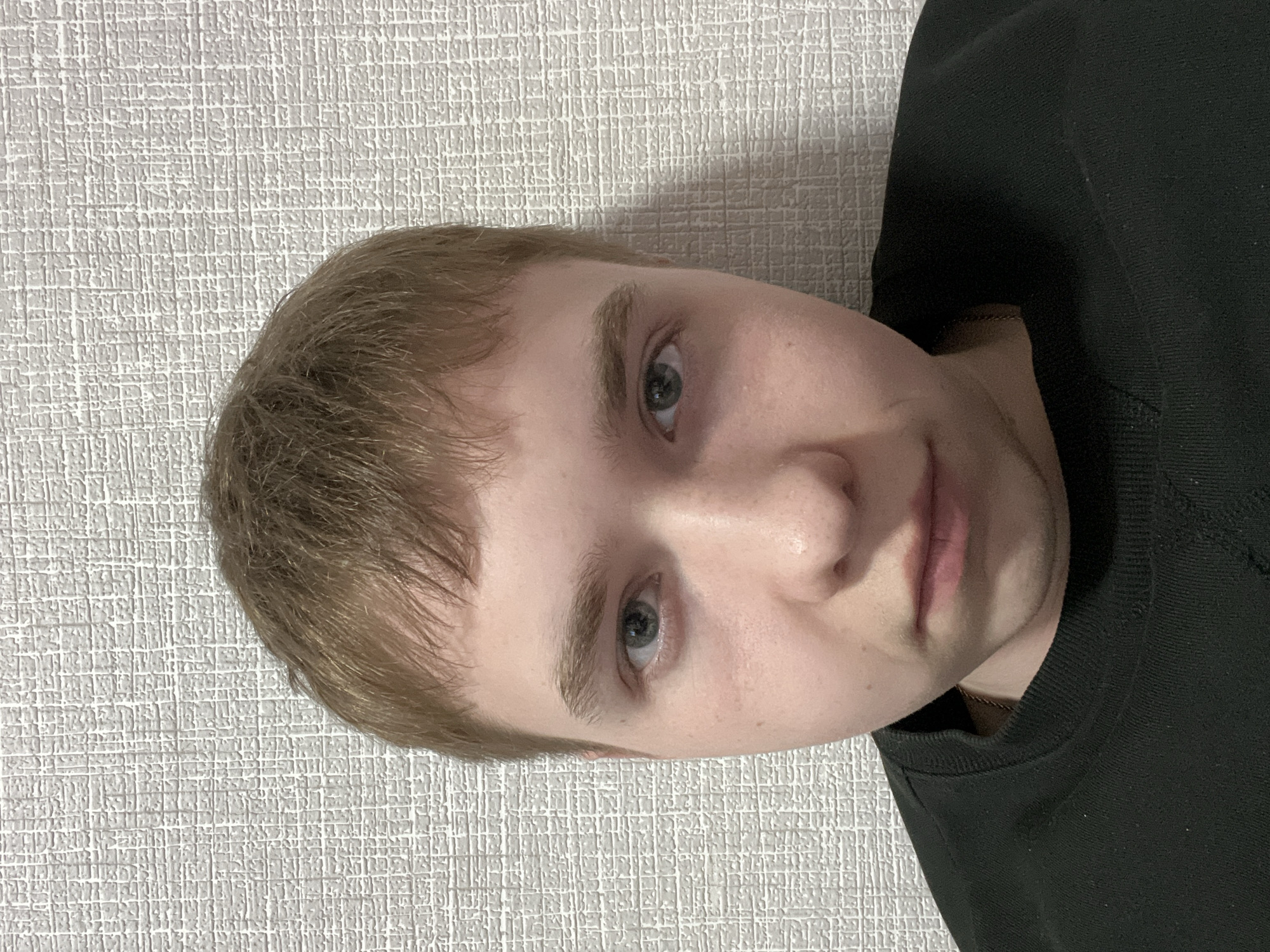 An indoor portrait features a young person, likely male, with fair skin, light brown, slightly wavy hair, and striking blue-grey eyes. The person is looking directly at the camera with a neutral, calm expression, featuring slightly parted lips. They are wearing a dark, possibly black or dark grey, long-sleeved shirt or sweatshirt, with a thin, gold-colored chain visible around their neck. The image is captured at a slight angle, with the person's head tilted.
The background consists of a light off-white or light grey wall, which displays a distinct, finely textured grid-like pattern. The lighting appears soft and even, typical of indoor conditions, with no harsh shadows, suggesting either diffused daylight or artificial room lighting. No specific time of day or weather conditions can be discerned. The setting is a straightforward portrait against a simple, textured wall. There is no visible text in the image. Given the provided context, the location is Perm, Russia.