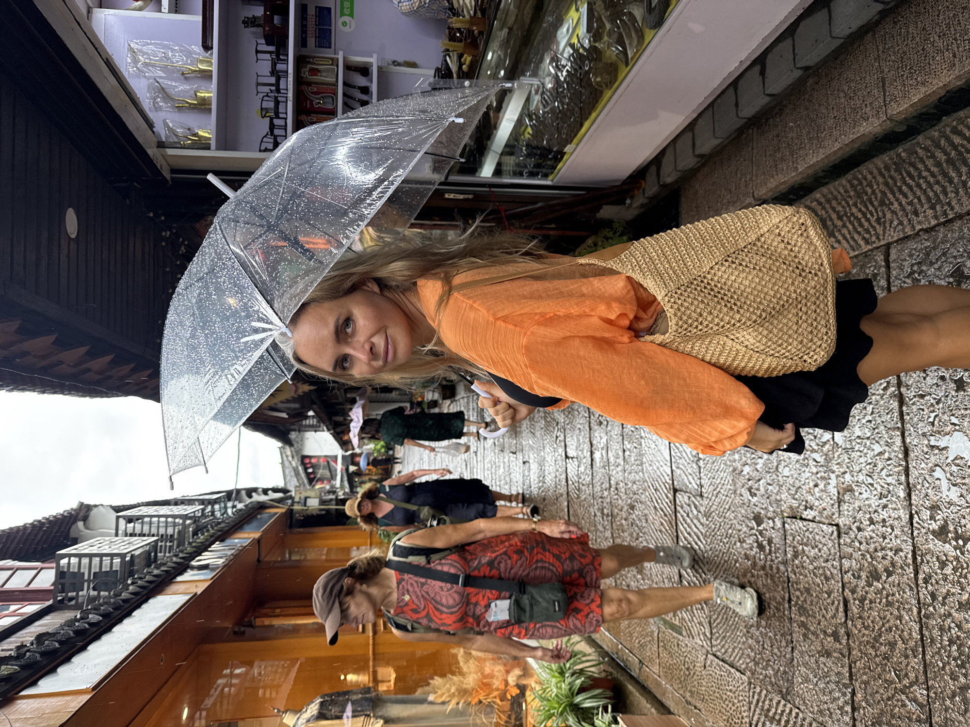 A woman holding a clear umbrella with water droplets on it stands on a cobblestone street in Qingpu District, China. She is looking back at the camera and smiling. She is wearing an orange blouse and a black skirt, and carrying a straw bag over her shoulder. The street is lined with traditional Chinese buildings, some with orange awnings. Other people are walking in the background, some also holding umbrellas. The ground is wet, indicating it is raining.