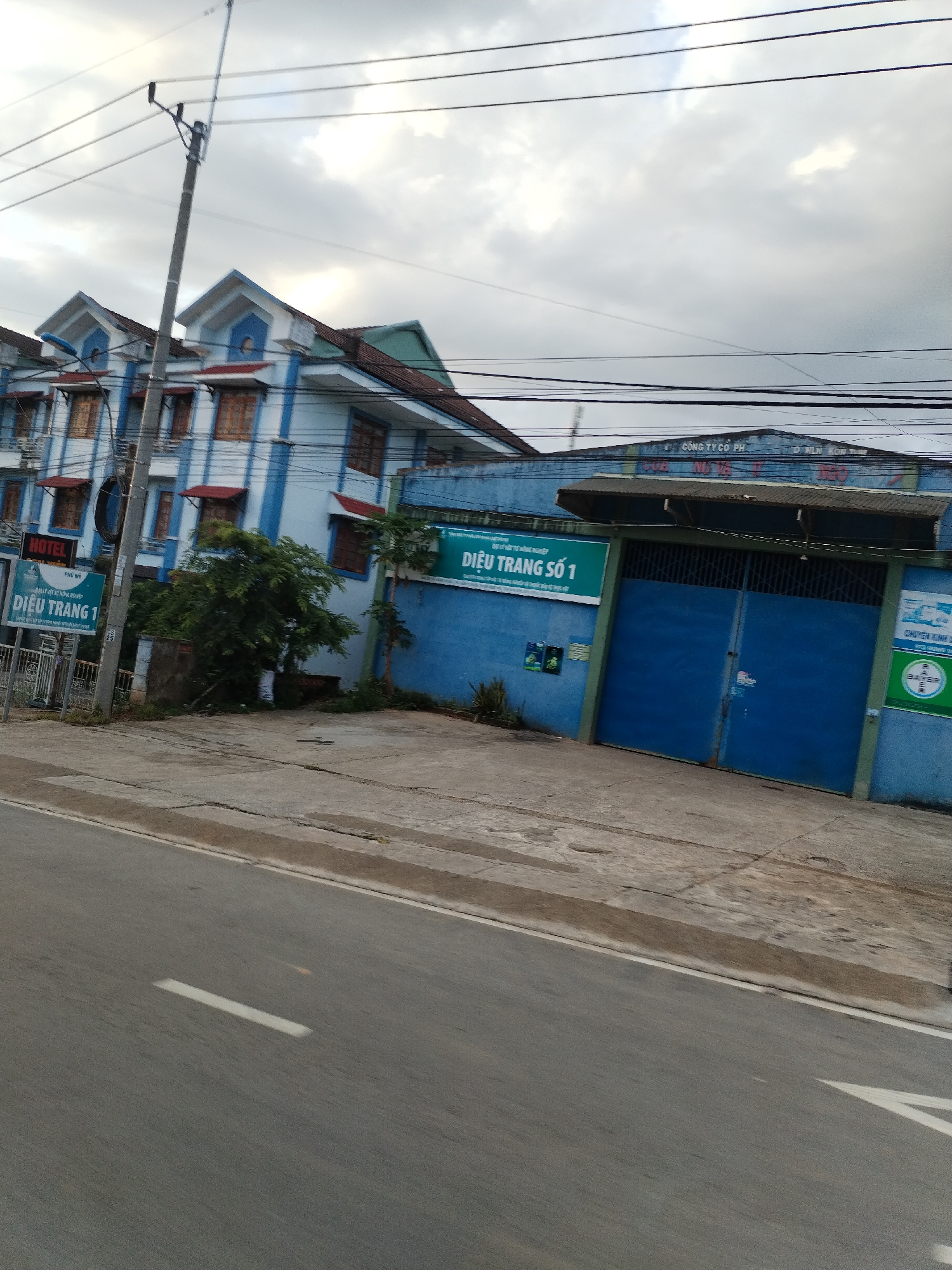 The image shows a street scene in Đăk Xú, Vietnam. On the left is a three-story hotel building painted light blue with dark blue trim, red awnings over the windows, and a sign that reads "HOTEL DIỀU TRANG 1". There is a utility pole to the left of the building with wires overhead.
To the right of the hotel is a large blue industrial building with large blue doors. A sign on the building reads "DIỆU TRANG SỐ 1" and another sign has the Bayer logo. A concrete road is in the foreground with painted white lane markings. The sky is overcast.