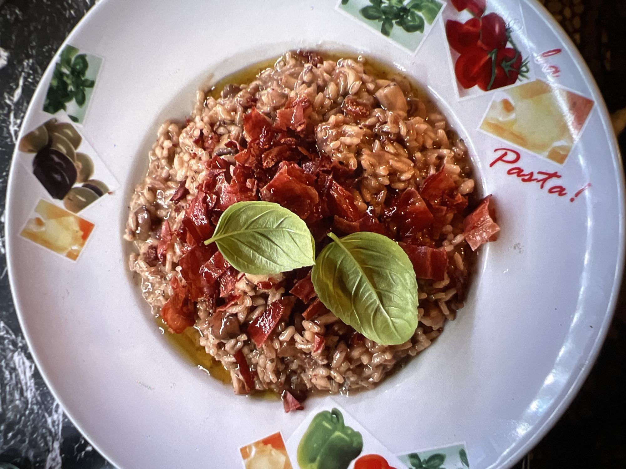 A close-up, top-down shot captures a white plate holding a portion of risotto. The risotto is a creamy, light brown color and is topped with crispy, crumbled pieces of what appears to be bacon or prosciutto, along with two fresh green basil leaves. The rim of the plate is decorated with small, square images of vegetables and herbs, including olives, peppers, tomatoes, and basil. The word "Pasta!" is written in red cursive script on the lower right portion of the plate, and below it, a small heart symbol is also present in red. The plate rests on a dark, marbled surface. The overall setting suggests a restaurant in Verona, Italy, likely a trattoria or a restaurant specializing in Italian cuisine, given the Italian script and imagery on the plate. There are no people or actions depicted.