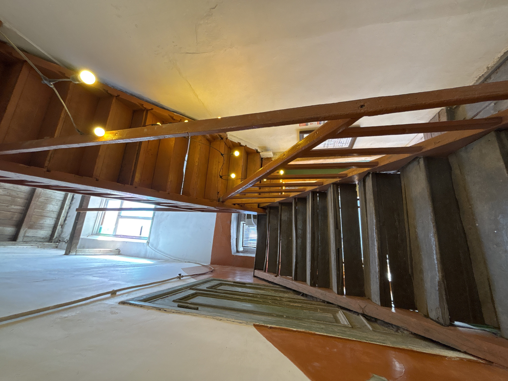 This image captures a narrow, indoor wooden staircase in Yerevan, Armenia. The staircase, made of dark wood, is worn and appears aged, with visible gaps between some of the treads. A wooden handrail, painted a reddish-brown, runs along the right side of the stairs. String lights with yellow bulbs are draped along the banister and the underside of the staircase, providing illumination. The walls flanking the stairs are painted in contrasting colors: the left wall has an upper section of white and a lower section of vibrant orange, while the right wall is predominantly white. To the left of the stairs, there is a distressed, light green wooden door with intricate paneling. At the top of the staircase, a small window with multiple panes is visible, letting in natural light. The ceiling above appears to be made of unfinished wooden planks. The overall impression is of a rustic and possibly historic interior space.