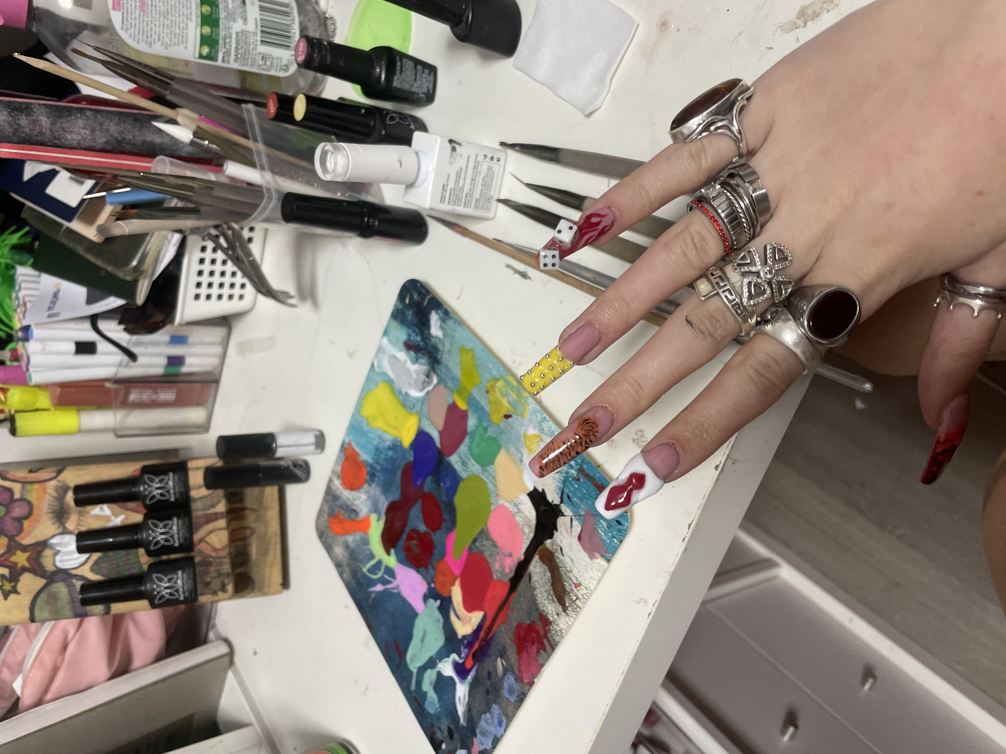 The image shows a cluttered workspace with nail art supplies. A hand with multiple rings and intricately designed nails is positioned above a palette of nail polish colors. Various nail art tools, including brushes and small bottles of nail polish, are scattered around the hand and palette. The image provides no contextual information to determine if the image was taken in Raanana, Israel.