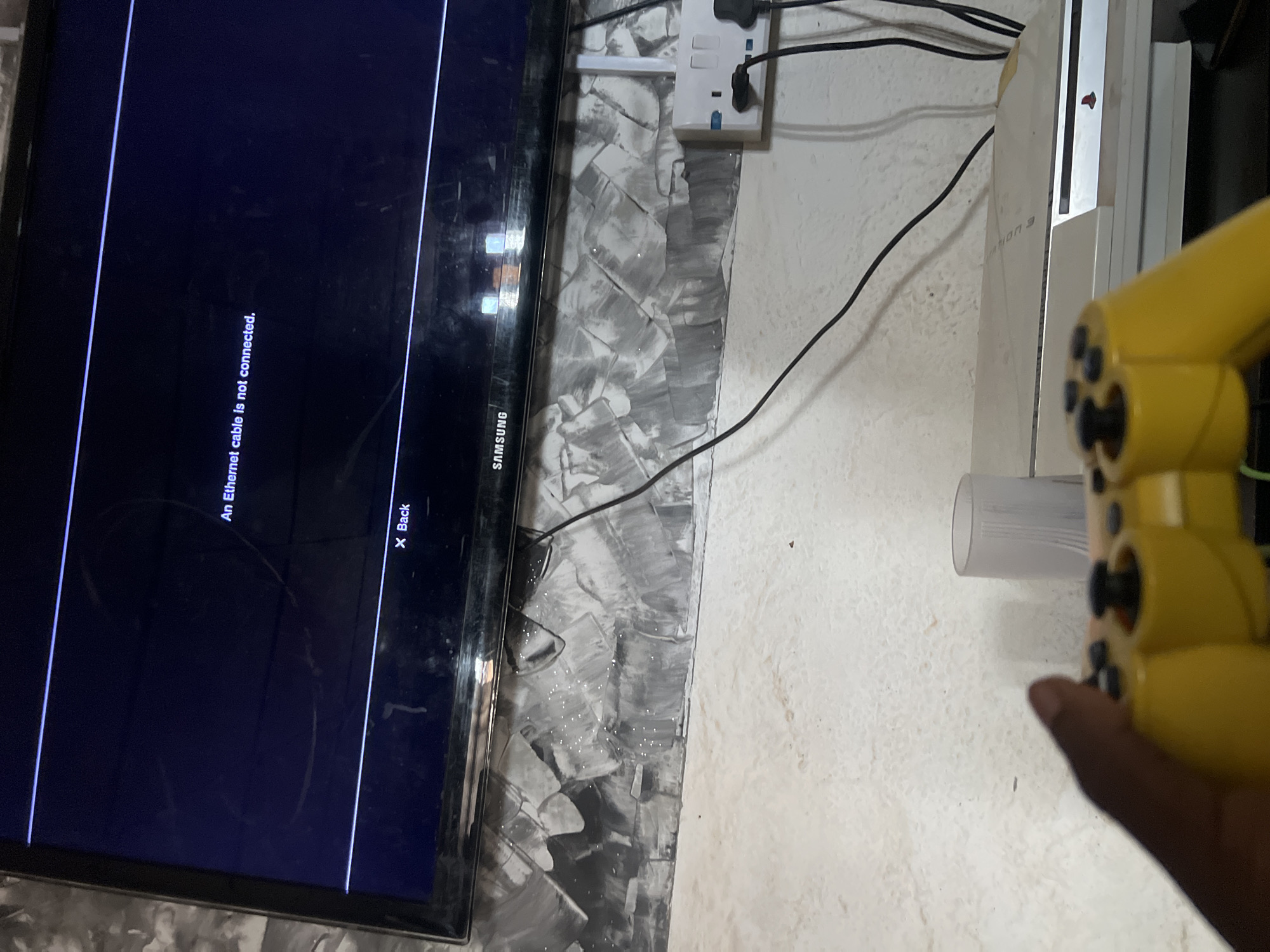 The image displays a close-up, low-angle shot of a television screen showing an error message: "An Ethernet cable is not connected." Below the television is a white PlayStation 3 console, stacked on top of another item. A yellow video game controller is partially visible in the foreground, held by a person whose hand is also in view. The background consists of a wall with a textured, gray and white patterned wallpaper, and a white electrical outlet with two black plugs inserted. The lighting appears to be ambient, coming from the screen and an unseen source. The scene is set indoors, possibly in a living room or entertainment area. The location is identified as Durimi, Nigeria.