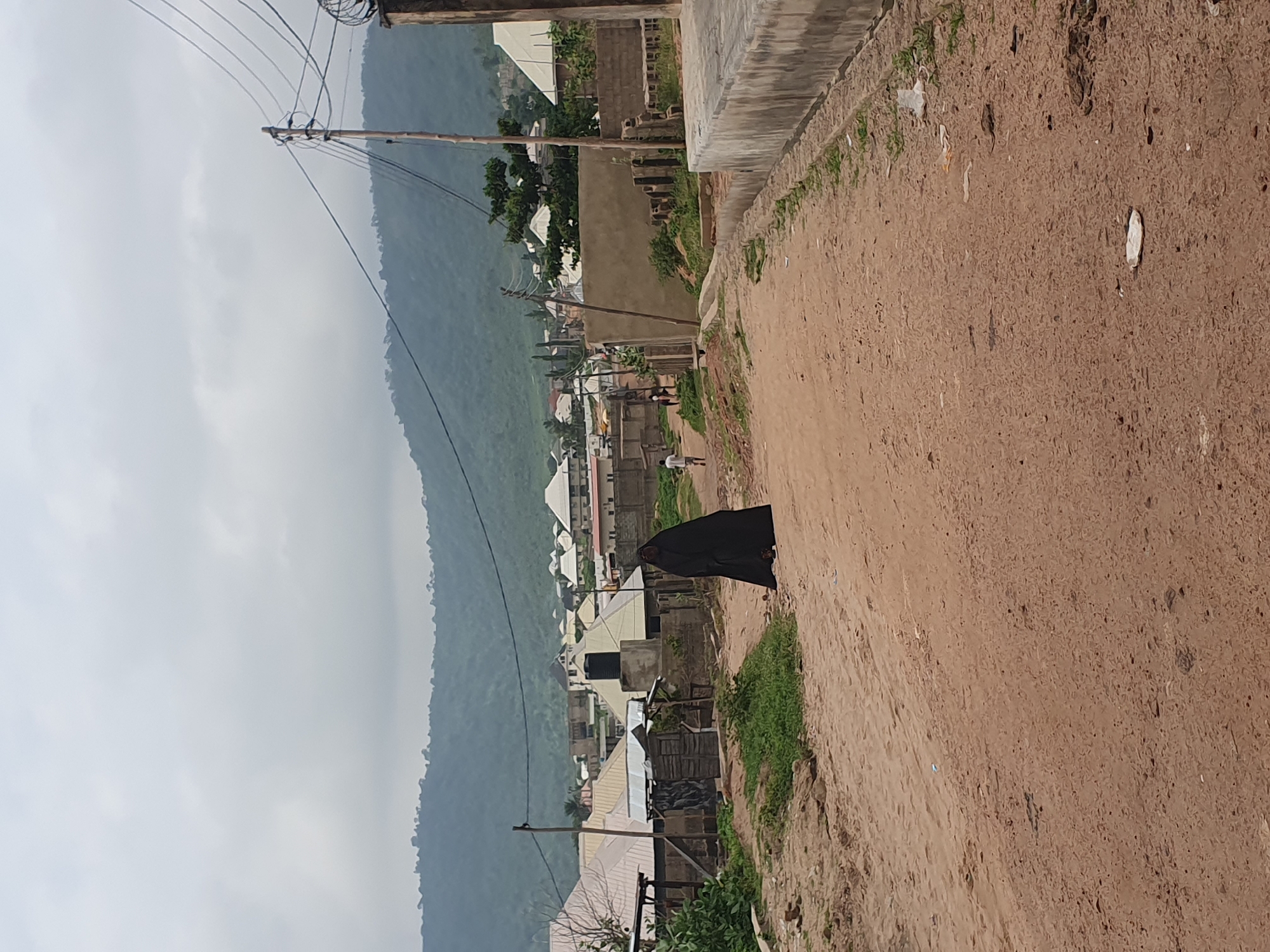 A person in a black abaya walks on a dirt road in Lokoja, Nigeria. In the background, there are houses with corrugated iron roofs, some of which are white. A large black water tank is visible on top of one of the buildings. Power lines and a utility pole are present in the scene. The road is unpaved and dusty, with some green vegetation on the sides. In the distance, a hill with scattered trees can be seen under a cloudy sky. Another person, wearing a white shirt, walks further down the road in the opposite direction. The scene depicts a residential area in Lokoja.