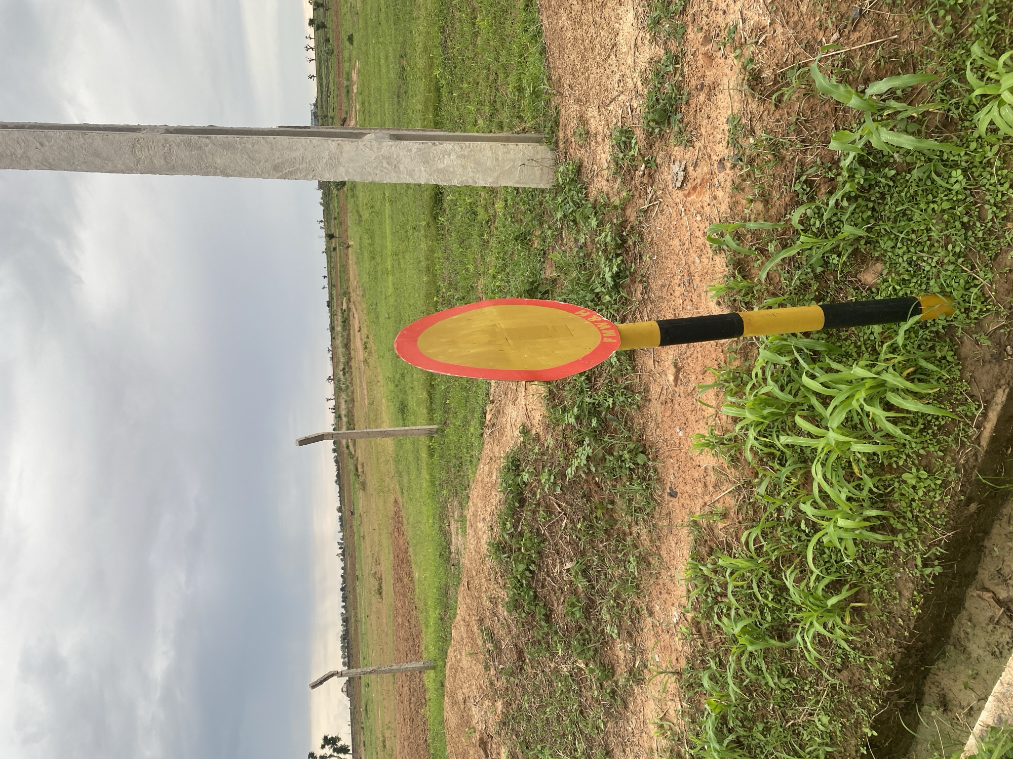 The image depicts an outdoor scene in Dumnga, Nigeria, characterized by an overcast sky suggesting it might be a cloudy day or late afternoon. The foreground is dominated by a broken traffic barrier, painted with alternating black and yellow stripes, with an oval red and yellow sign attached. The sign has some text that is partially visible, reading "H&M" in red letters. Behind the barrier, a concrete pole stands vertically, likely part of a construction or infrastructure project.
The setting is a rural or undeveloped area, with a mixture of dry, sandy soil and patches of green grass and vegetation. In the background, a vast expanse of green fields stretches out, with some scattered trees and possibly some distant structures or farmsteads. The fields appear to be agricultural land, with signs of cultivation or natural growth. The overall impression is one of an open, somewhat uncultivated landscape under a subdued sky. There are no people visible in the image, and the focus is on the natural environment and the man-made barrier and pole. The scene does not convey a specific event, but rather a static view of the location.