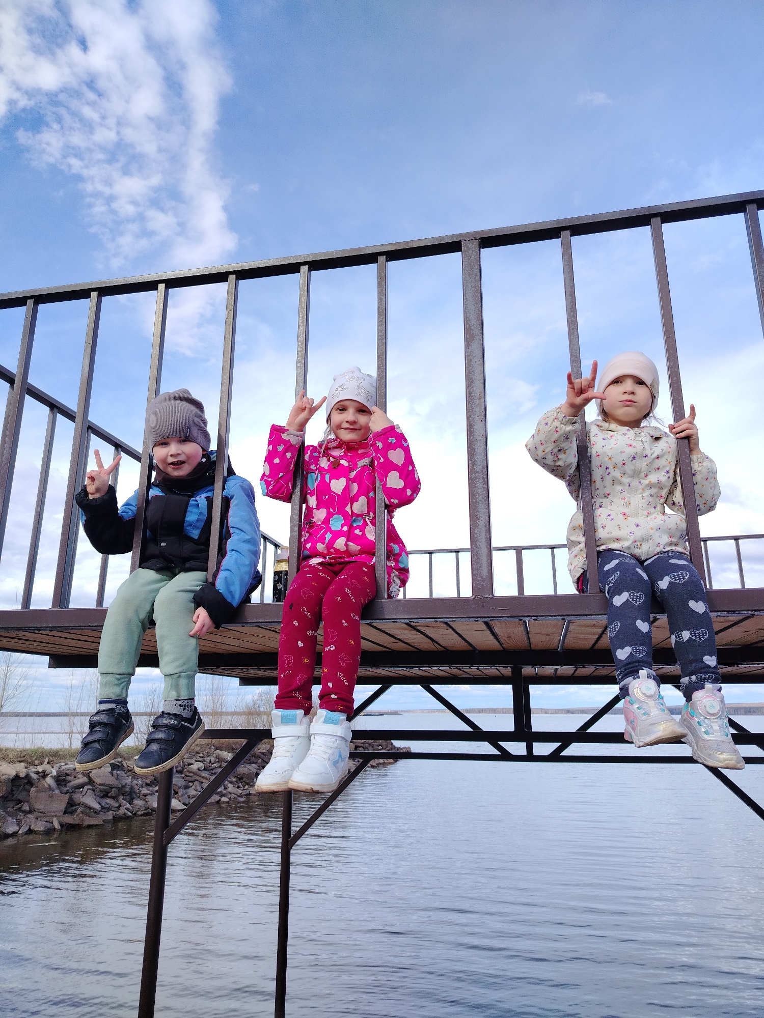 Three young children are playfully posing on a wooden platform with metal railings, overlooking a large body of water in Chaikovsky, Russia. The boy on the left, wearing a grey beanie, blue and black jacket, and light green pants, flashes a peace sign with both hands. The girl in the middle, dressed in a white beanie, a vibrant pink heart-patterned jacket, and red pants, makes an "I love you" hand gesture with both hands. The girl on the right, with a light beige beanie and a floral-patterned jacket over dark blue heart-patterned pants, also forms the "I love you" sign with both hands. All three children are smiling brightly at the camera, their expressions reflecting joy and youthful energy.
Their legs dangle freely above the calm, grey-blue water, which shows reflections of the overcast sky. A rocky shoreline is visible on the left, leading to a distant landmass across the expanse of water. The overhead sky is light blue with scattered white clouds, indicating a bright but possibly cool day, aligning with their jackets and beanies. The platform appears to be part of a pier or observation deck. Visible text on the boy's shoes includes "CLASSIC" and "GOODIE PARIS." The scene captures a happy outdoor moment during daylight.