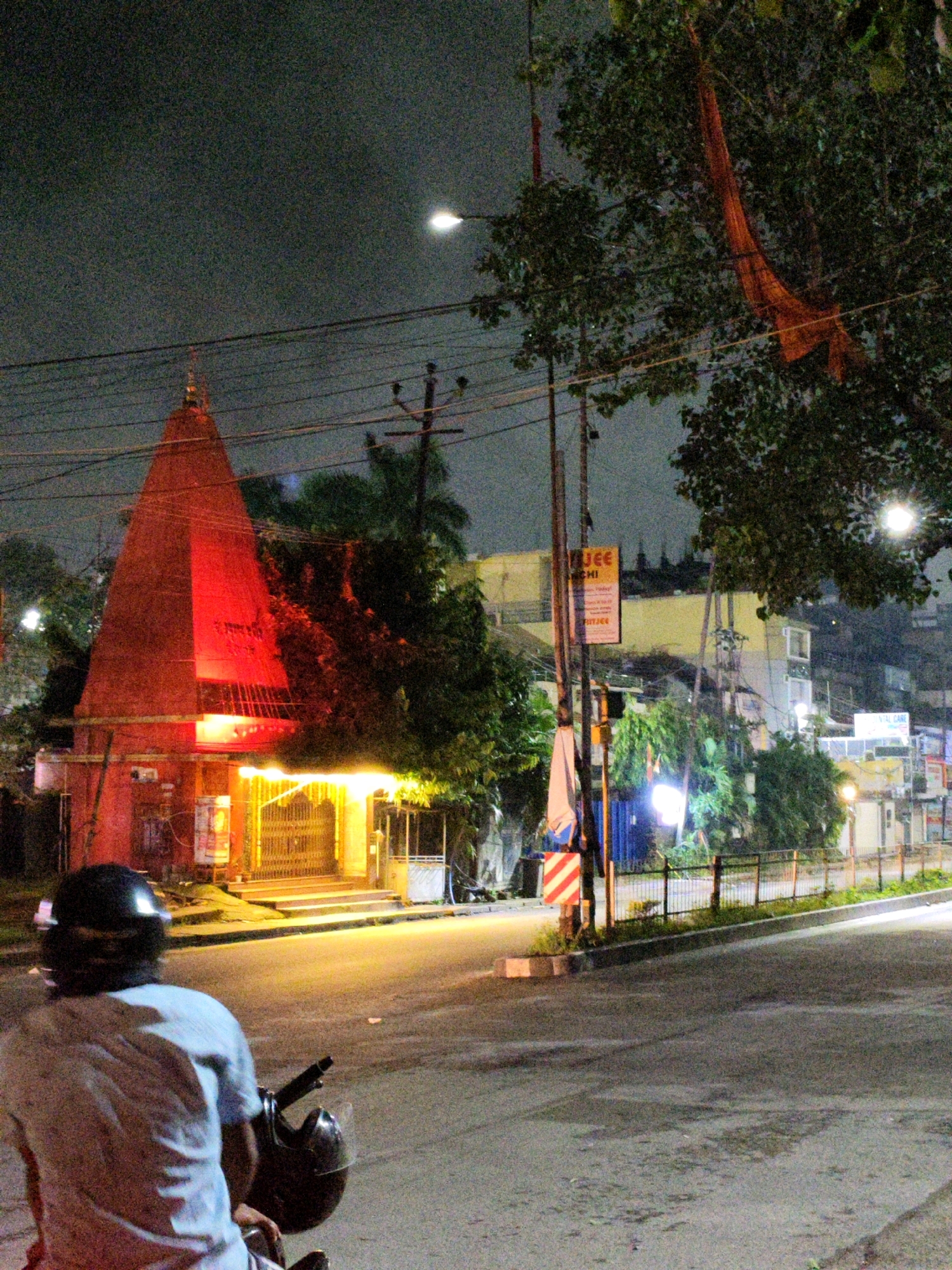 The image captures a street scene at night in Ranchi, India. In the foreground, a person wearing a light-colored shirt and a black helmet is seated on a motorcycle, viewed from behind. Adjacent to the motorcycle is a dark-colored helmet. The motorcycle is parked on an asphalt road, which is illuminated by streetlights and the lights emanating from a nearby building.
Dominating the left side of the image is a tall, conical structure, appearing to be a temple or shrine, illuminated with bright red light. It has a prominent entrance with a large, ornate gate that is brightly lit with yellow lights, suggesting an active place of worship. The architecture of the temple appears to be traditional, with a pointed spire.
Behind the temple, trees are visible, with dense foliage contributing to the nocturnal ambiance. A large, orange cloth or banner is draped over a tree branch in the upper right portion of the image. Further in the background, several buildings of varying heights and architectural styles can be seen, some with illuminated windows. A street lamp with a bright light is visible in the upper portion of the frame. Signs, one of which appears to be a billboard, are also present on some of the buildings. The overall environment suggests a moderately urban setting with elements of religious significance, characteristic of areas within Ranchi. The presence of streetlights and the illumination of the temple indicate that this is a public area, likely on a main road. The scene is peaceful and quiet, typical of a city at night.